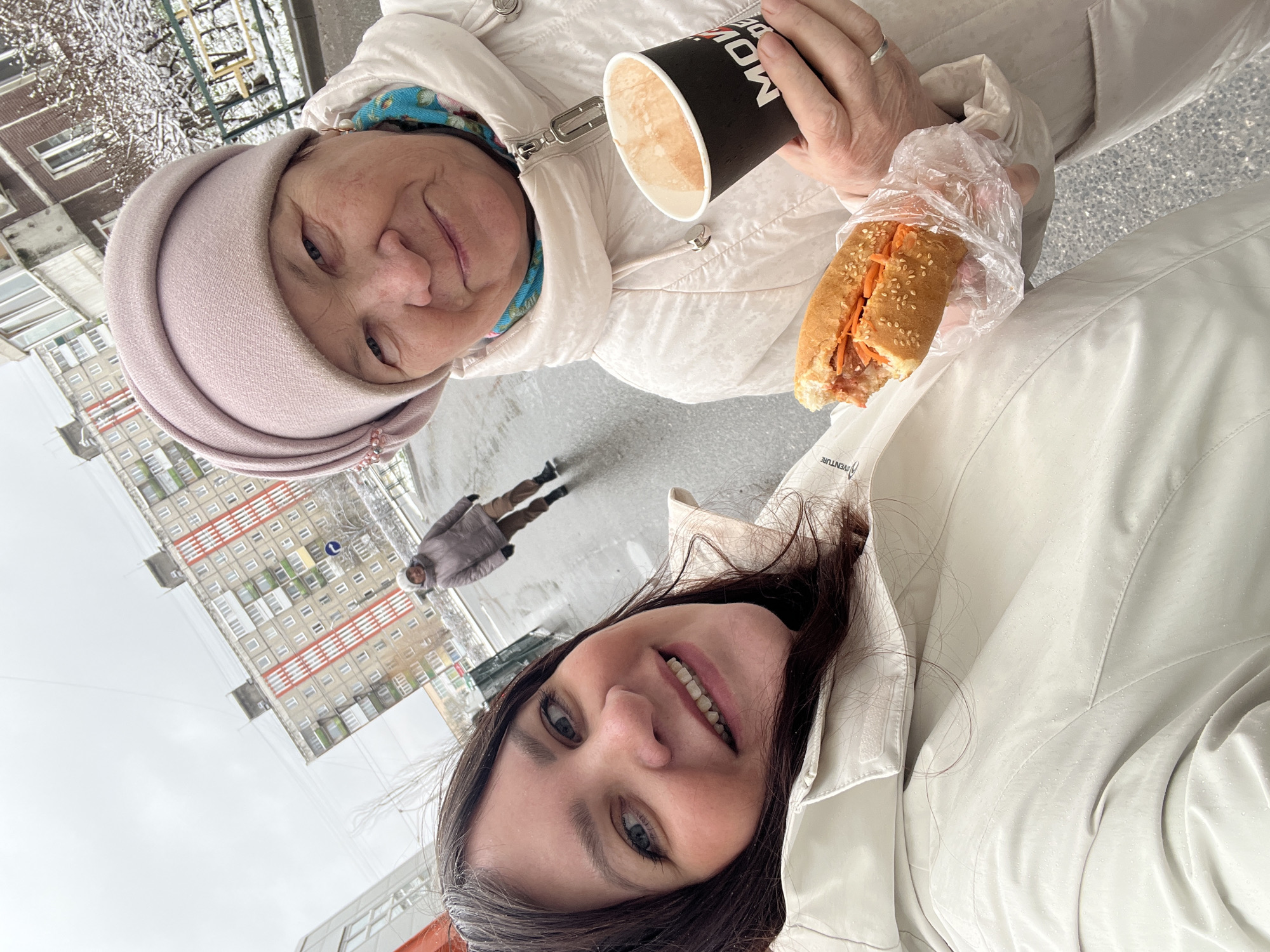 A rotated selfie-style outdoor photograph captures two women smiling in an urban setting during winter in Perm, Russia.
In the foreground, an elderly woman with a gentle smile wears a light-colored winter jacket and a light pink head covering. She holds a black cup with a beverage, partially displaying white text like "MOBA" or "MOV...", and a partially eaten hot dog or sandwich. The hot dog, encased in clear plastic, features a sesame seed bun and a filling of what appears to be meat and shredded orange vegetables.
Below her, a younger woman with dark hair, also in a light-colored winter jacket, smiles broadly, revealing her teeth. Visible water droplets on her jacket suggest recent rain or snow. She appears to be holding the camera for the selfie.
The background depicts multi-story residential buildings, typical of Soviet-era architecture, under an overcast sky. Snow dusts bare tree branches, reinforcing the winter season. A third person, a male figure, is seen walking in the middle ground on a street or sidewalk. The scene conveys a friendly, casual outing on a cold, damp winter day.