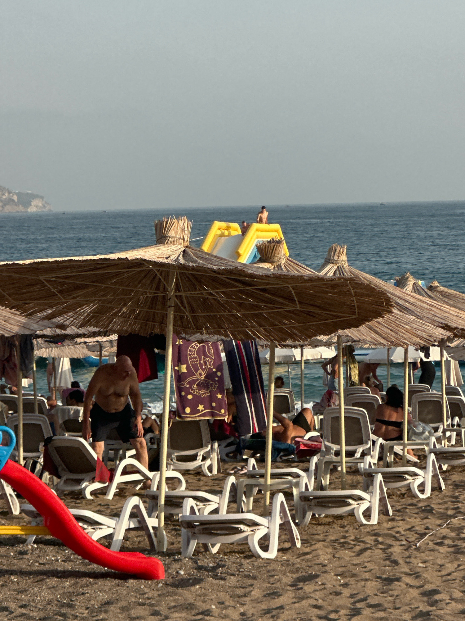 The image depicts a crowded beach scene, likely in Boreti, Montenegro, given the mountainous coastline visible in the background. The foreground is dominated by numerous thatched umbrellas providing shade for rows of white lounge chairs. A red children's slide is positioned on the left side of the frame, with yellow accents. Several individuals are visible on the beach, including a man in black shorts standing under an umbrella and appearing to be adjusting his clothing. Other beachgoers are seated in lounge chairs, some facing the water, while others are partially obscured. In the background, the blue ocean stretches to the horizon, with a few small boats visible. A large inflatable yellow water slide is set up in the water, with at least two people on it. The overall atmosphere is one of leisure and recreation on a sunny day.