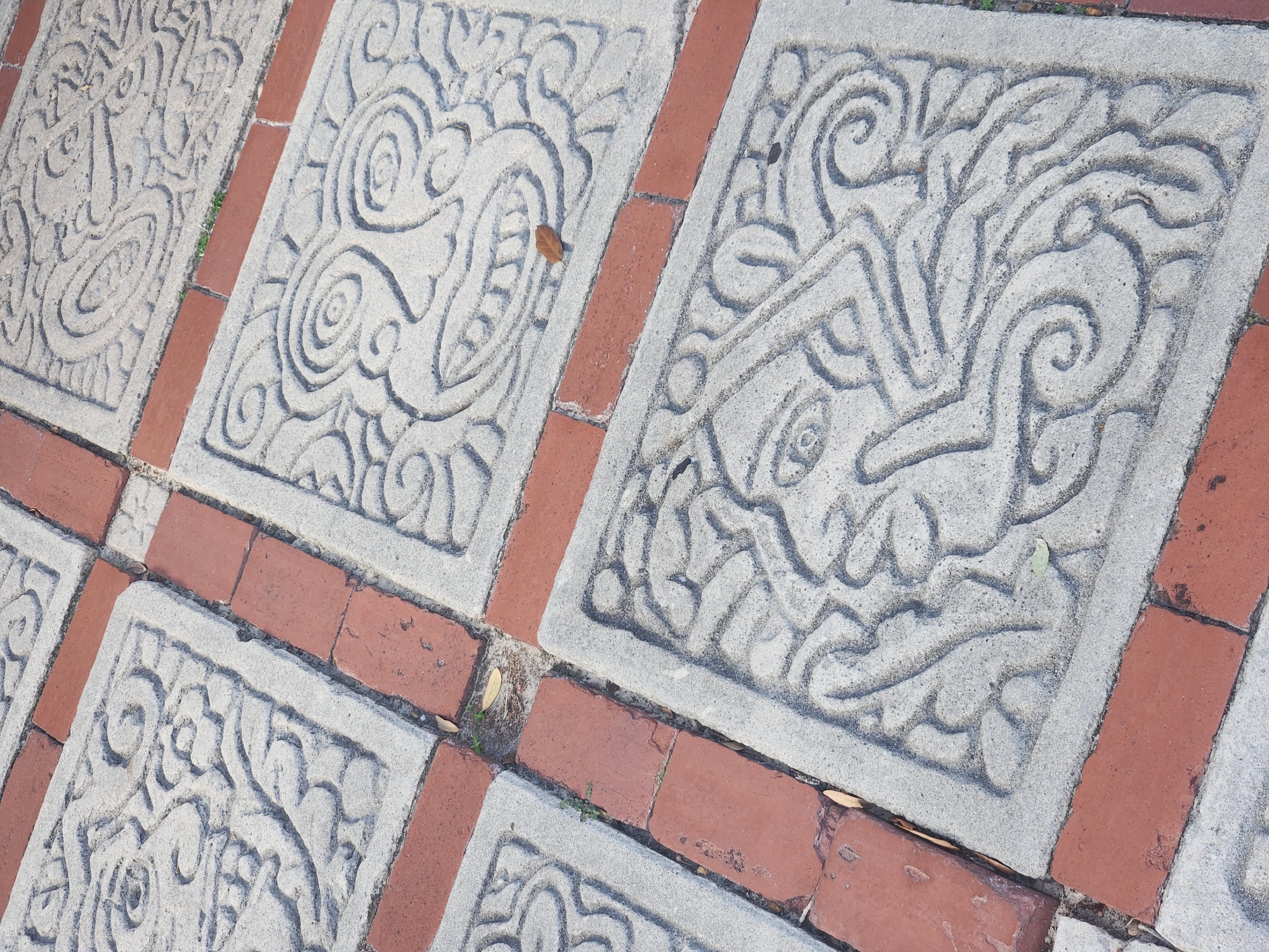 This is a close-up, high-angle shot of a paved pathway featuring decorative square concrete tiles interspersed with red bricks. The concrete tiles are intricately carved with abstract, stylized faces or masks, featuring swirling patterns and bold lines. The bricks are laid in a pattern that complements the concrete tiles. The pathway appears to be outdoors, possibly in a park or public space. The lighting suggests it is daytime, with sunlight casting shadows and highlighting the textures of the materials. There are a few fallen leaves and some sparse green vegetation visible in the crevices between the tiles and bricks. The overall impression is one of detailed craftsmanship and a possibly cultural or artistic design choice for the paving. The location in the city of Maitland, United States of America, is not directly evident from the image content itself, but the style of the carvings might suggest a specific cultural influence if known.
