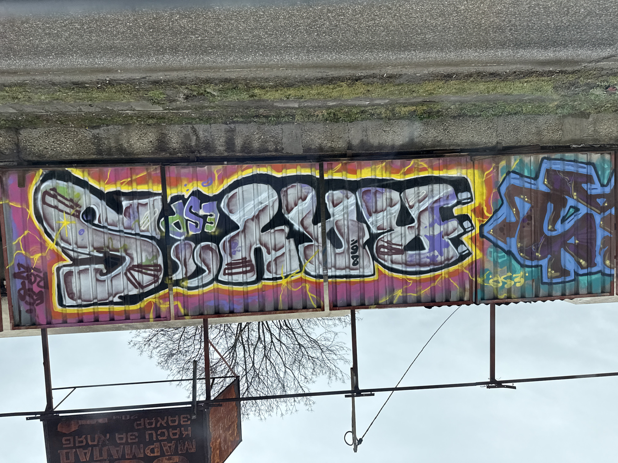 The image shows graffiti on a corrugated metal fence. The graffiti on the left spells out "SPICE" in large, stylized letters, outlined in black with a yellow and pink background. To the right is another graffiti tag in blue and brown with a green outline. Above the fence is a concrete ledge or sidewalk. Below the fence are bare tree branches against a cloudy sky, and part of a rusty sign with Cyrillic writing. The location is likely in Sofia, Bulgaria due to the Cyrillic text.