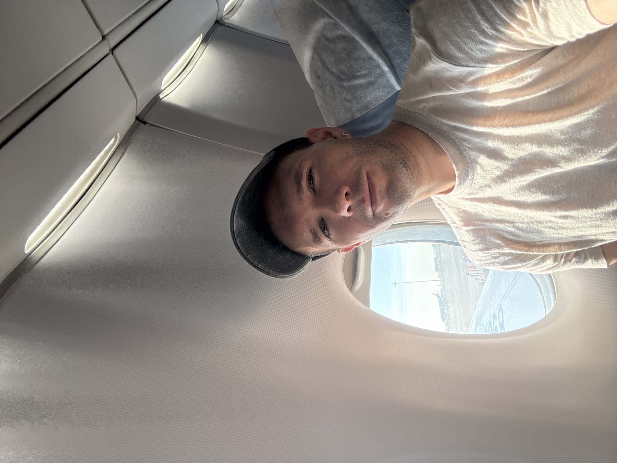 A man wearing a grey baseball cap and a light-colored t-shirt is seated on an airplane. He is looking towards the camera, with a slight smile. The airplane cabin is visible, with the ceiling panels and overhead lights illuminated by the sun. A window next to the man shows a view of an airport tarmac with an airplane wing in the foreground and other aircraft and a control tower in the distance. The image appears to have been taken at Sheremetyevo International Airport, located in Khimki, Russia, as evidenced by the airport scenery visible through the window.