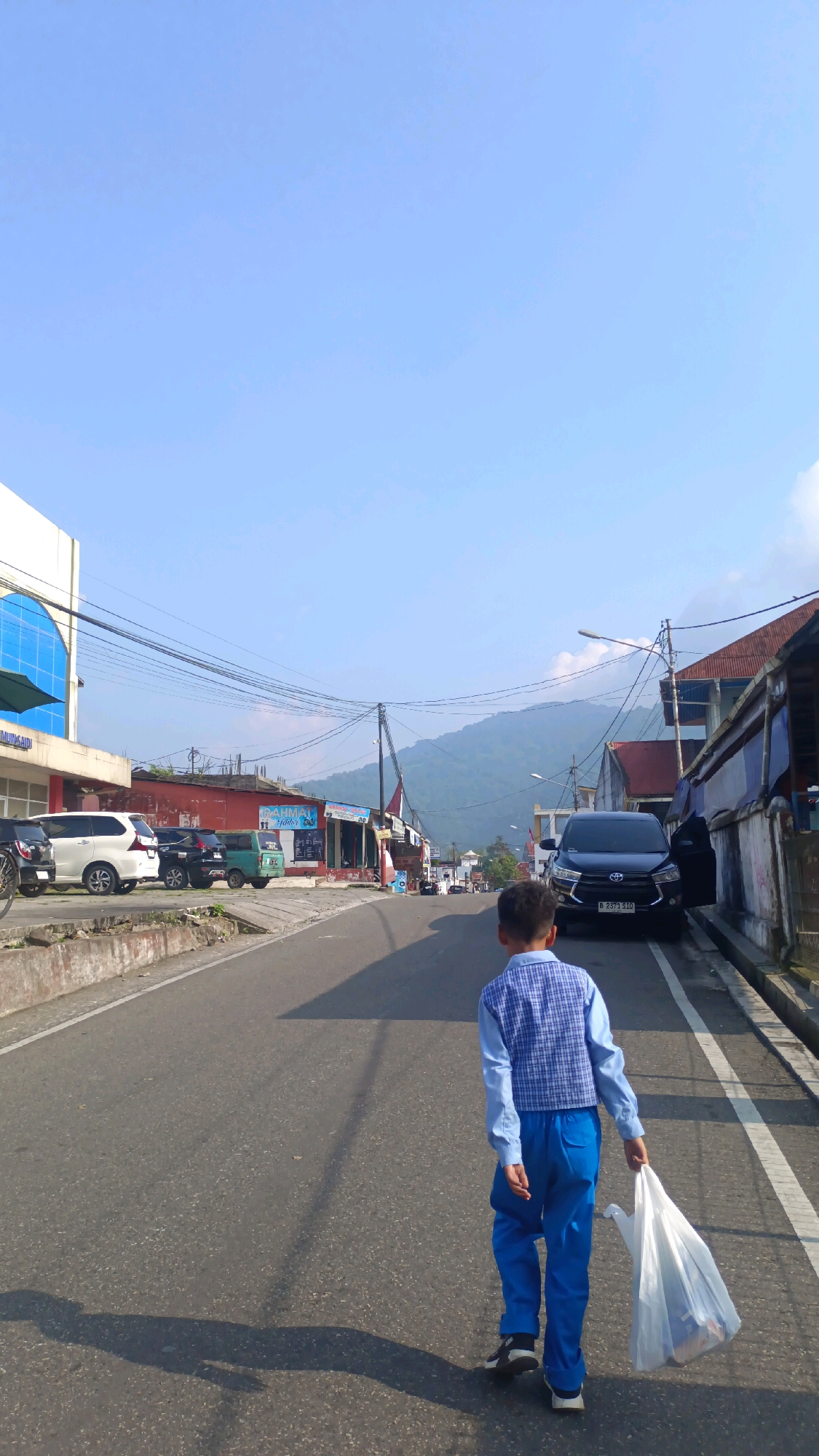 A young boy, wearing a blue plaid vest over a light blue long-sleeved shirt and blue pants, walks away from the camera down a paved road in Padang Panjang, Indonesia. He carries a white plastic bag in his right hand. The boy is in the right foreground of the image. The road is lined with buildings on both sides. On the left, a modern building with blue accents and a curved window is visible, followed by a row of shops. On the right, older buildings with corrugated metal roofs and weathered facades are present. Several cars are parked along the left side of the road, and a black SUV is parked on the right. The sky is clear and blue, and a hazy mountain range can be seen in the distance. The scene appears to be illuminated by bright daylight, casting shadows on the road. The image captures a typical street scene in a town with a mix of modern and traditional architecture.