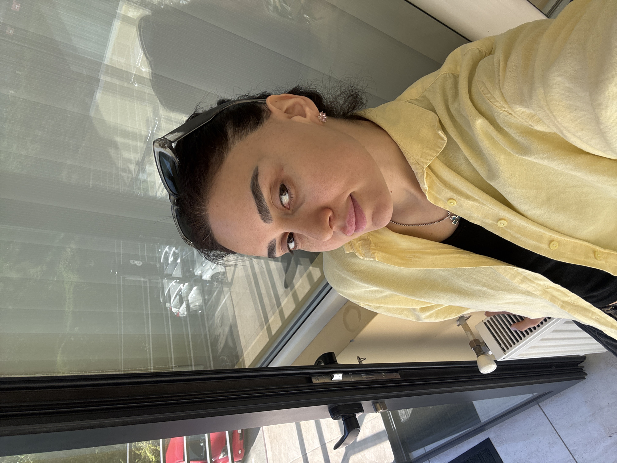 A medium shot captures a woman indoors. She is wearing a yellow shirt over a black top, and sunglasses are perched on her head. She appears to be looking towards the camera. Outside the window, a scene is visible, likely reflecting a street in Tbilisi, Georgia, given the red vehicle and visible surroundings suggestive of an urban environment. The room contains a window with blinds, a black frame, and what appears to be a radiator or heating unit near the bottom of the frame. The floor is tiled. The image is rotated somewhat counterclockwise.