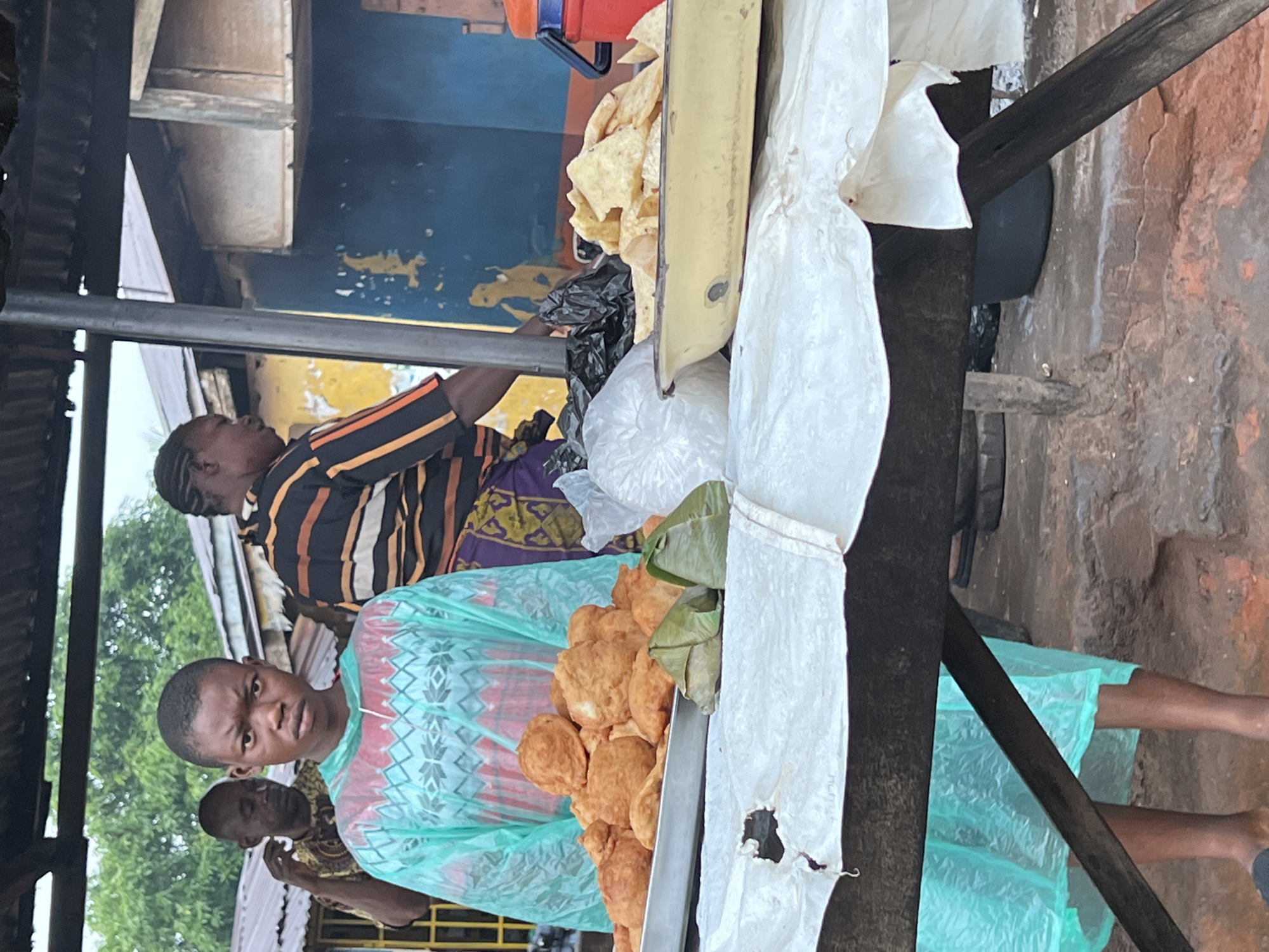 In Nkpor, Nigeria, a street vendor stands behind a display of fried food, possibly puff-puff or similar fritters, arranged in a metal tray. The food is golden brown and some pieces are wrapped in green leaves. The vendor is a young person wearing a teal-colored garment with a diamond pattern. Behind them, another person wearing a striped shirt with black, orange, and yellow lines is visible, along with another person partially obscured. The scene appears to be an outdoor market or roadside stall, with a rustic wooden structure supporting the display. There is a blue and yellow painted wall in the background, and a black plastic bag is present on the stall. The overall atmosphere suggests a typical day in a busy market. The lighting is diffused, indicating it could be daytime, with overcast weather perhaps contributing to the soft light. The ground appears to be dirt or rough concrete.