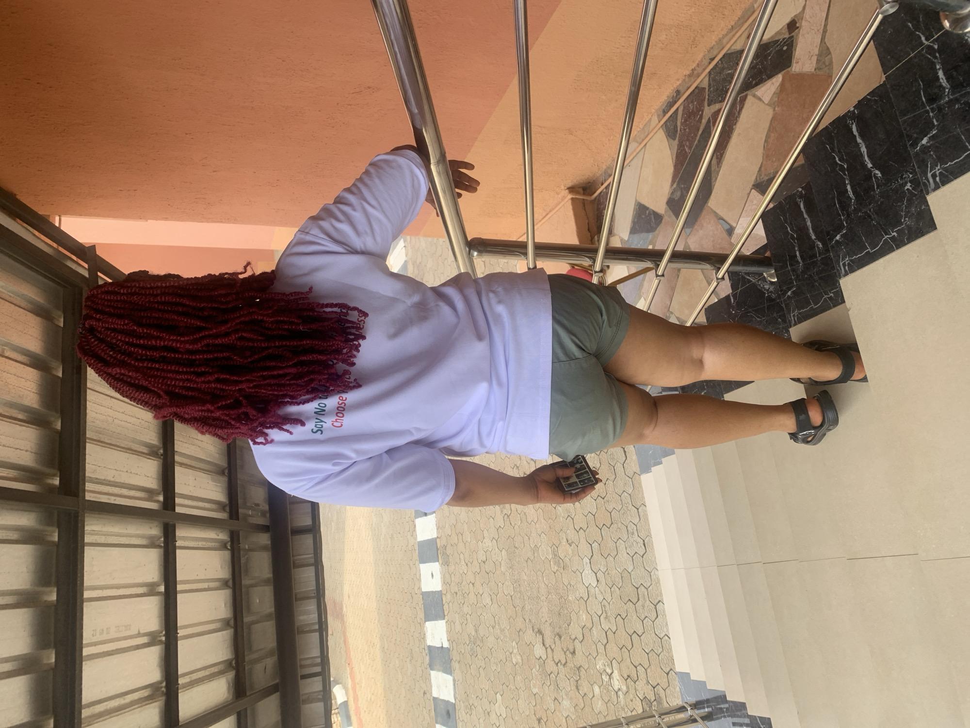 A person with maroon braided hair is standing on a landing of a staircase in Gura Top, Nigeria. They are wearing a white long-sleeve shirt that says "Say No to Rape" in red lettering, and olive green shorts, and black sandals. The person is holding a small black object in their right hand. The staircase has stainless steel railings. The flooring around the landing consists of tan tiles in a geometric pattern and black marble tiles with white veining. A metal grid-like structure is visible to the left. A portion of a tan wall is visible above the staircase.
