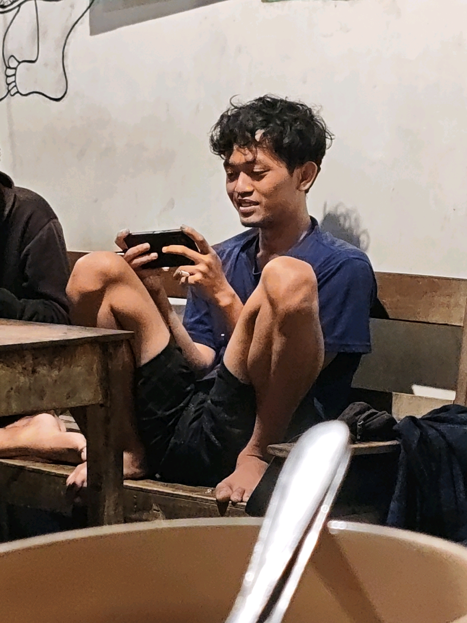 In the city of Ngasem, Indonesia, a young man with curly black hair is seated on a wooden bench, engrossed in playing a mobile game on his smartphone. He is wearing a blue t-shirt and dark shorts. His legs are bent, and his feet are bare. To his left, another person is partially visible, wearing a dark jacket, seated at a wooden table. The background features a white wall with a large, abstract drawing of a foot. The lighting suggests an indoor setting, possibly a cafe or a common area, with some shadows cast on the wall. In the foreground, a portion of a brown bowl and a shiny metallic object, possibly cutlery, are visible.