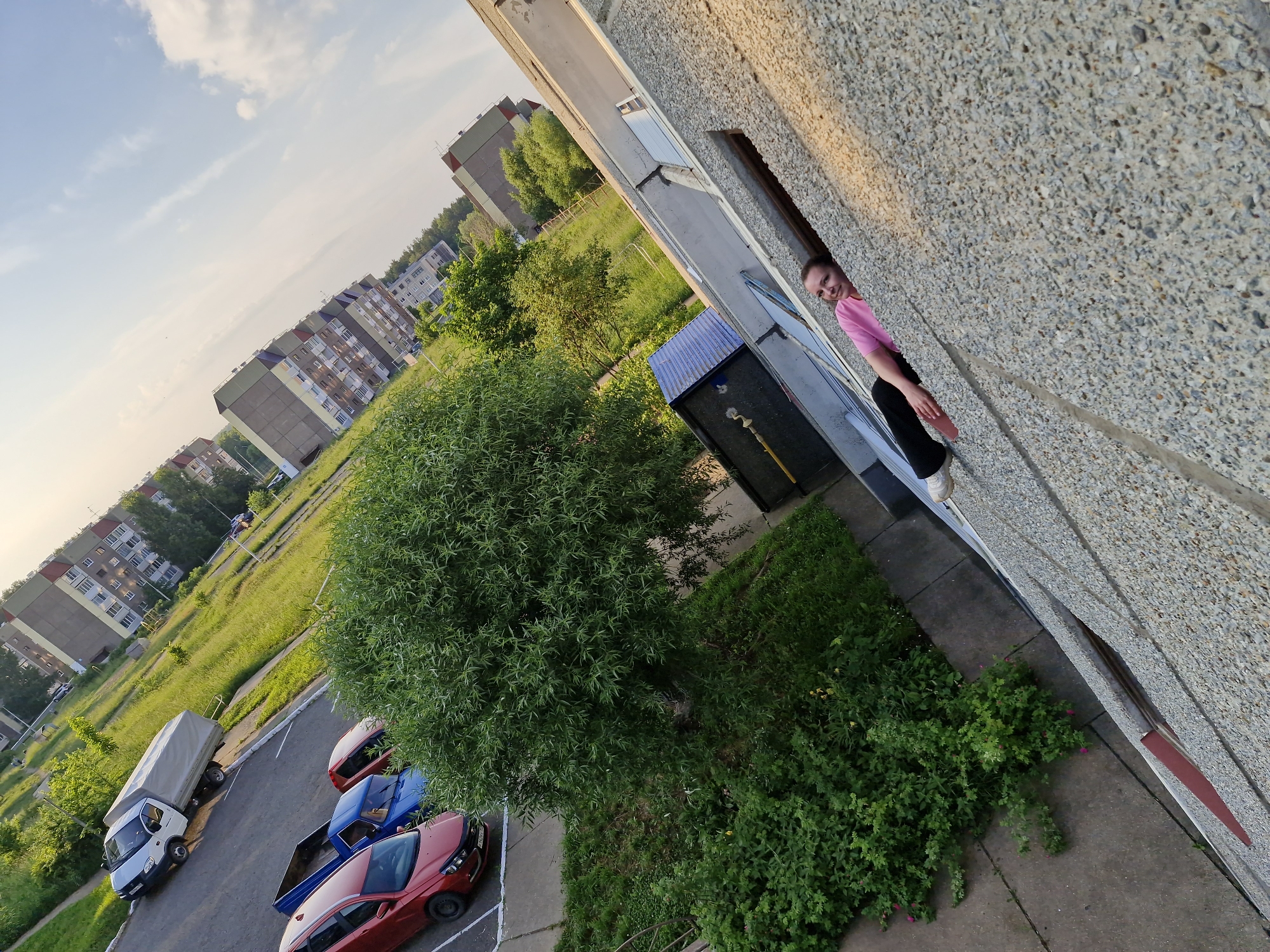 A young woman in a pink shirt and black pants is leaning out of a window on the third floor of a concrete apartment building, with her head and shoulders visible. The building has a rough, pebbled facade. Below the window, there is a blue and black utility shed. In the background, several cars are parked on an asphalt lot, including a white truck with a covered bed, a blue pickup truck, and red cars. The area is surrounded by green grass and trees, with several apartment buildings visible in the distance. The sky is blue with scattered clouds. The image is taken at an angle, giving a skewed perspective of the scene. The location is identified as Марковский, Russia.