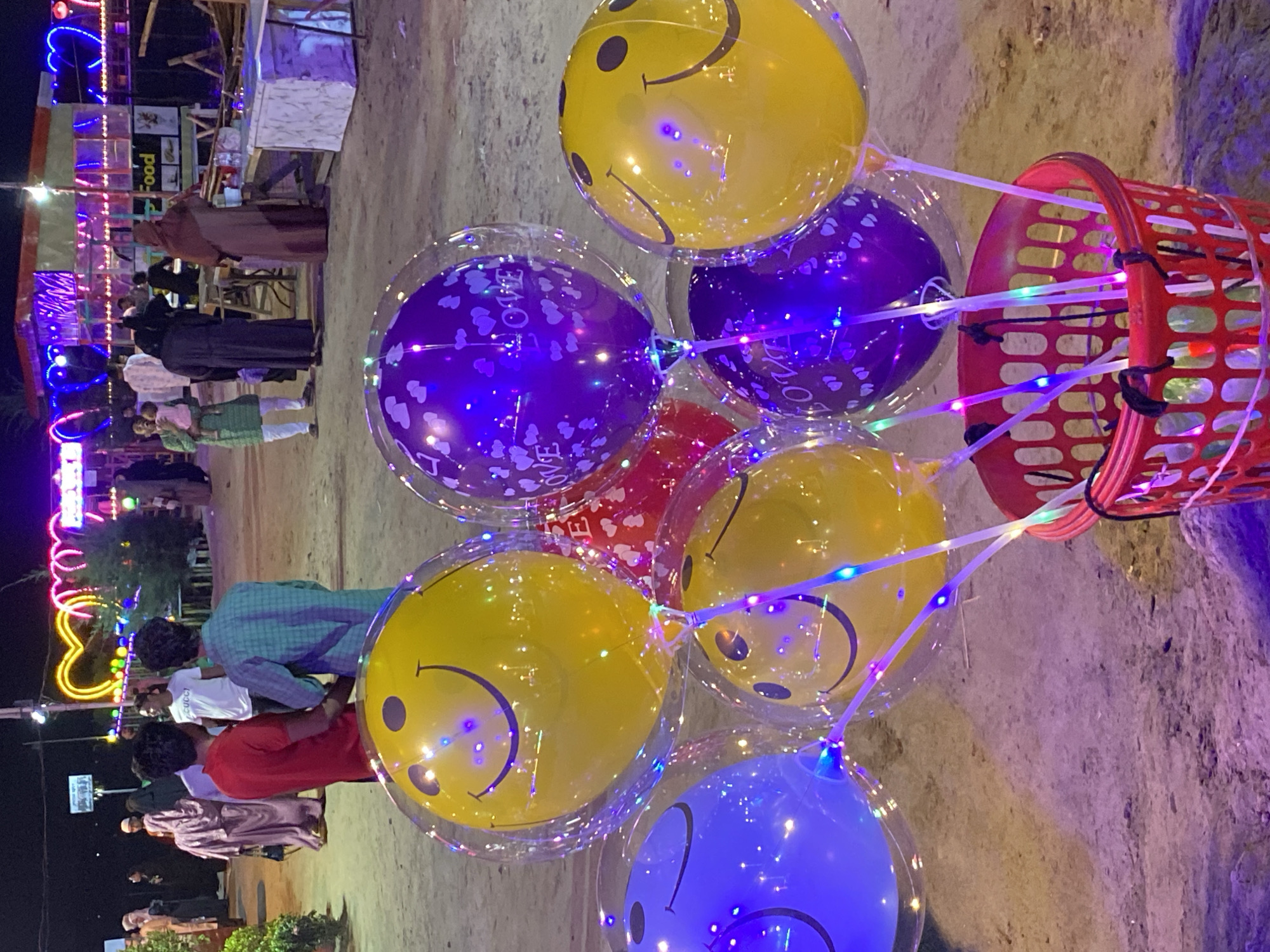 The image shows a street scene in Dhaka, Bangladesh. A red plastic basket containing a collection of illuminated balloons with smiley faces is prominent. The balloons are in varying colors, including yellow, purple, red, and blue, with decorative lights attached to the strings. People walk by in the background, some appearing to browse at food stalls. A building or structure is partially visible in the background, adorned with decorative lighting. The ground appears to be a dirt surface.