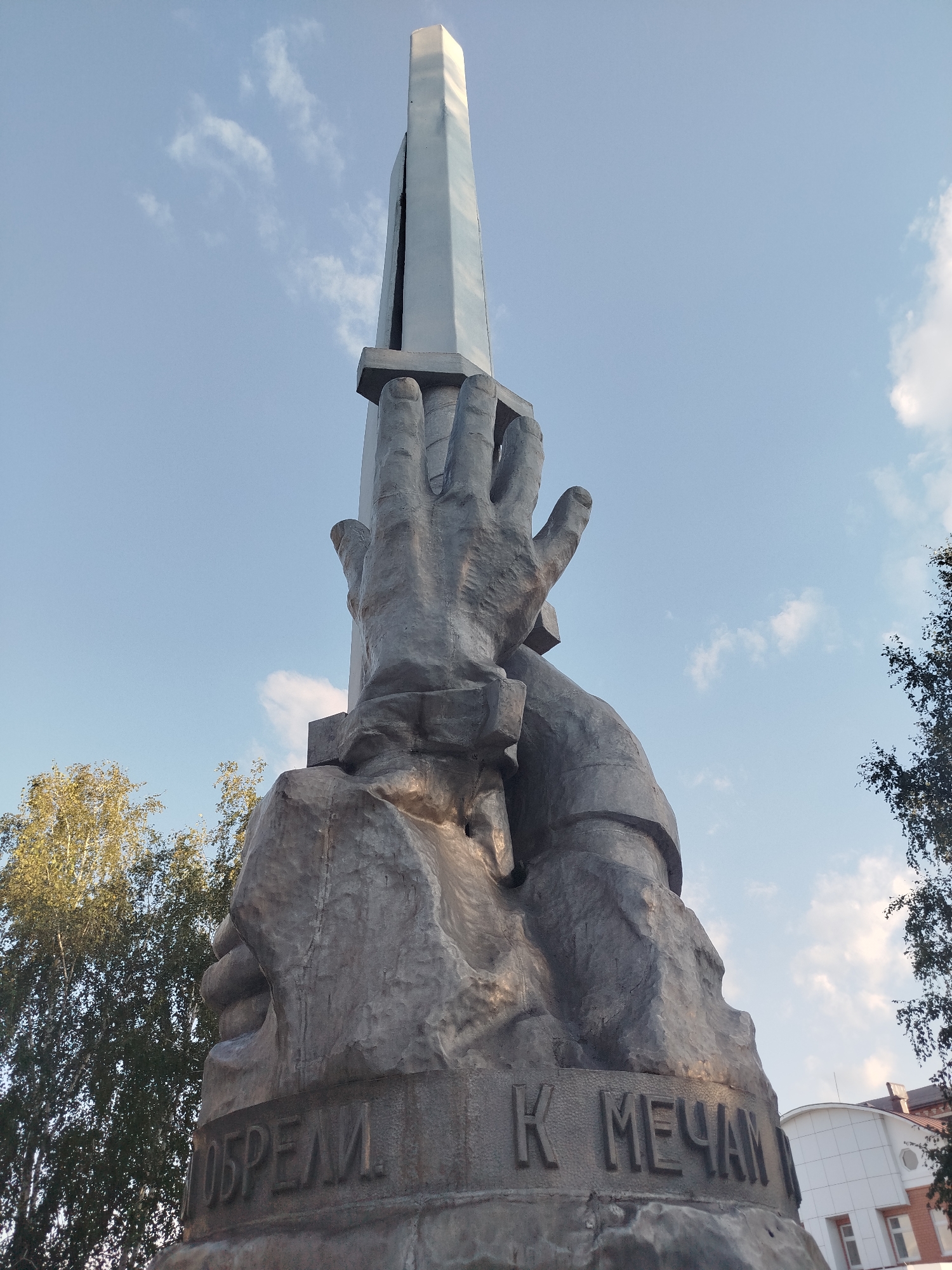The image shows a low-angle view of a large metal sculpture in Yalutorovsk, Russia, against a clear sky with scattered clouds. The sculpture depicts a pair of monumental hands reaching upwards, clasped around a sleek, metallic obelisk. The base of the sculpture is rough and textured, with carved Cyrillic lettering visible on the front. The letters appear to read "ДОБРЕЛИ К МЕЧТАМ", which translates to "Became to Dreams" or "Achieved Dreams". To the left of the sculpture, the tops of leafy trees are visible, and to the right, the upper portion of a modern building with white panels and windows can be seen. The overall impression is one of aspiration and achievement, with the sculpture reaching towards the sky.