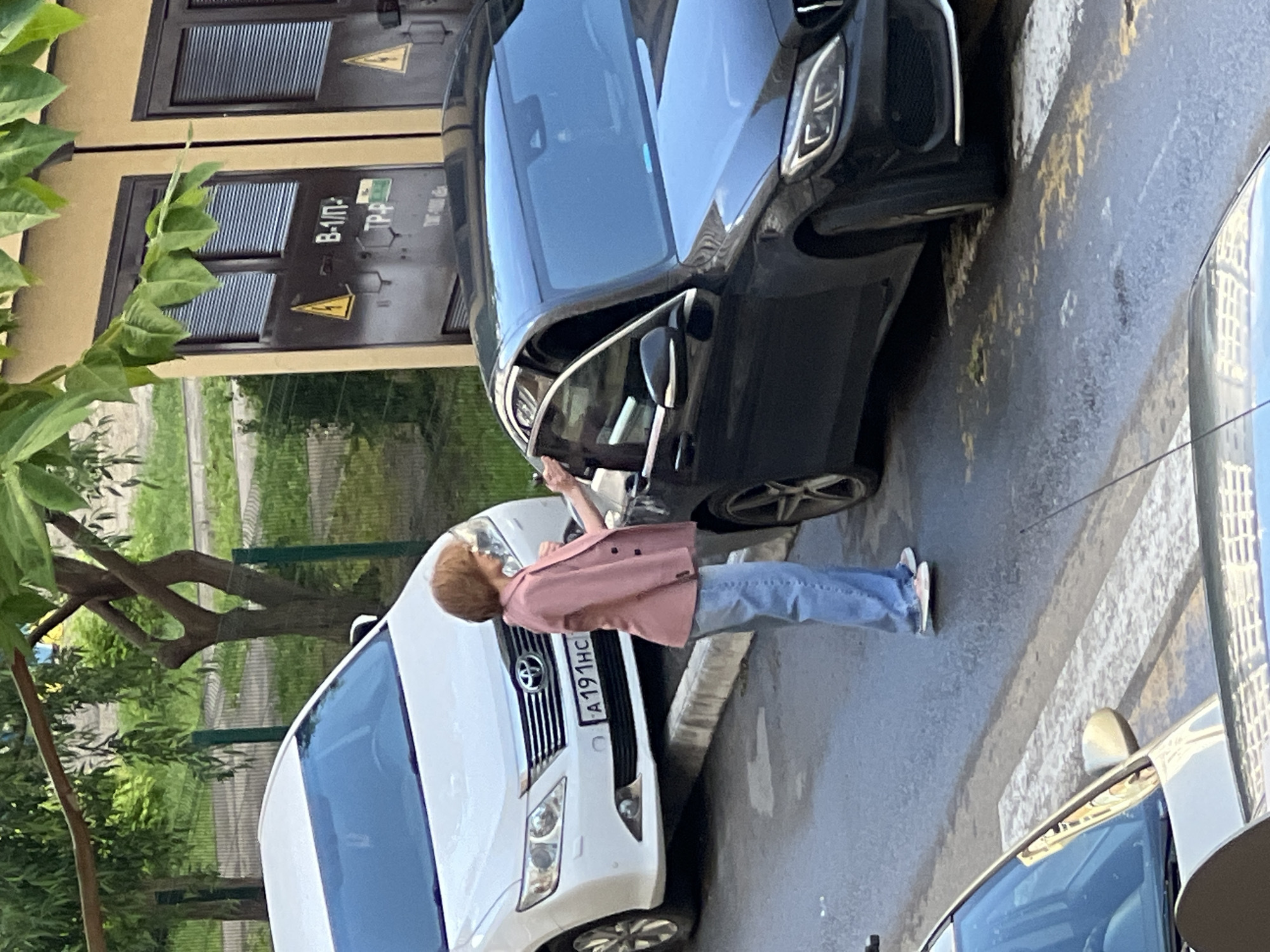 A woman in a pink blazer and blue jeans stands between a black Mercedes-Benz and a white Toyota SUV. The SUV has a license plate that reads "A191HC." The woman is reaching toward the Mercedes' side mirror with her right hand. A tree is on the left, and a utility box with the text "B-II-П" and a hazard symbol is on the wall behind the black Mercedes. The scene appears to be set on a city street in Krasnodar, Russia.