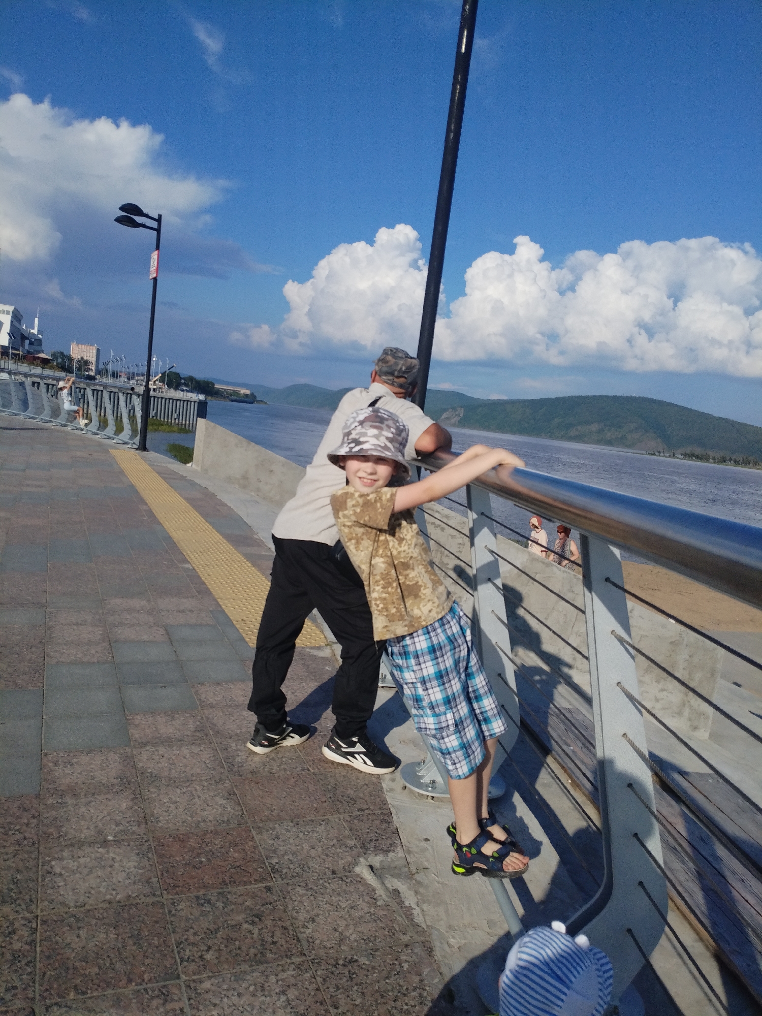 A young boy and an adult man stand on a paved promenade next to a body of water. The boy, in the foreground, leans on a metal railing and faces the camera, wearing a camouflage shirt, blue plaid shorts, and sandals. The adult man, behind the boy, is dressed in a light-colored shirt, black pants, and a camouflage hat, and looks out at the water. In the background, a river or lake stretches towards rolling green hills under a blue sky with scattered white clouds. Buildings and lampposts line the promenade, indicating an urban or riverside setting.