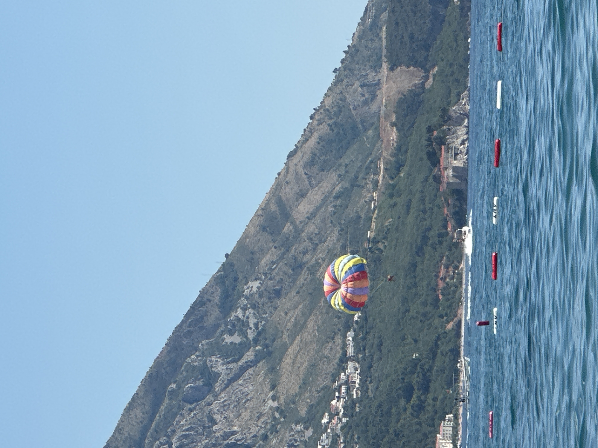 The image captures a parasailor being towed by a boat over the Adriatic Sea in front of the coastal town of Boreti, Montenegro. The parasail is colorful, with segments of red, yellow, green, blue, and purple. A person is visible hanging beneath the parasail. In the background, the mountainous landscape of Boreti rises steeply, dotted with buildings and lush green vegetation. The sea is a deep blue, with gentle waves and a line of buoys marking a swimming area. Several boats are visible on the water, including a speed boat and what appears to be a larger tourist vessel. The sky is a clear, bright blue, indicating a sunny day.