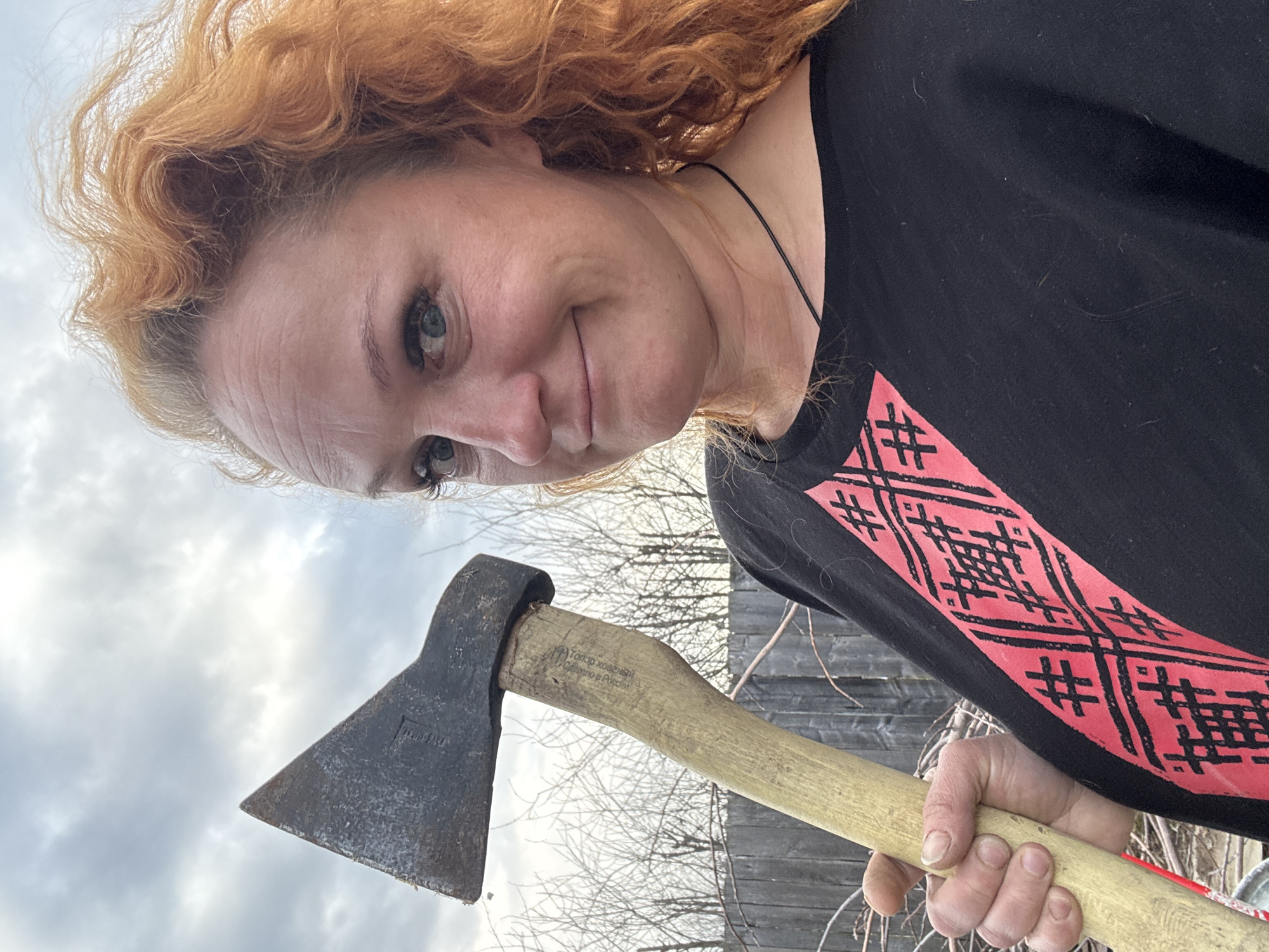 A close-up, angled portrait features a woman with vibrant, curly red hair and light eyes, looking directly at the camera with a subtle, almost mischievous smile. She wears a black long-sleeved shirt adorned with a distinctive red and black geometric pattern on the chest/sleeve area.
She holds a rustic axe, its wooden handle gripped in her hand, with the dark metal axe head positioned prominently towards the lower left of the frame. The axe head appears weathered and well-used.
Visible text on the axe includes "УРАЛ" (Ural) on the metal head, and the phrases "Топор кованый" (Forged axe) and "Сделано в России" (Made in Russia) clearly stamped into the wooden handle.
The setting is outdoors under an overcast or cloudy sky, providing soft, diffused lighting. In the background, bare tree branches and a weathered wooden fence or structure are visible, suggesting an autumnal or early spring day in an outdoor, possibly rural, environment. The overall impression is informal and suggests the woman is posing with a tool, perhaps in the context of outdoor work or leisure. The location is Chaikovsky, Russia.