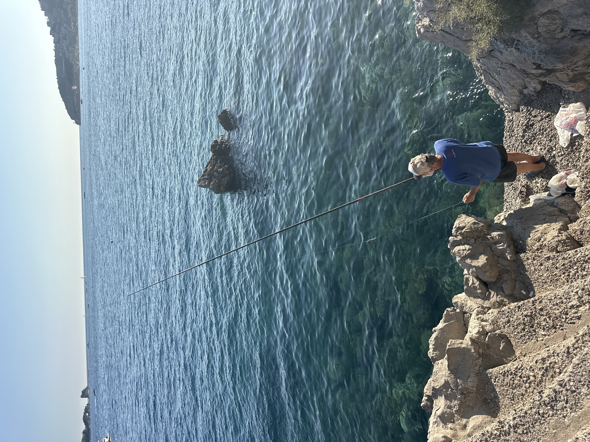 A man is fishing from a rocky coastline. He is wearing a blue t-shirt, dark shorts, and a baseball cap. His fishing rod is extended over the clear blue water, where a rocky outcropping is visible. The sea is calm, and the sky is clear and bright. In the distance, the coastline of Budva, Montenegro, can be seen with buildings and vegetation. There are two plastic bags on the ground near the man, one containing what appears to be fish.