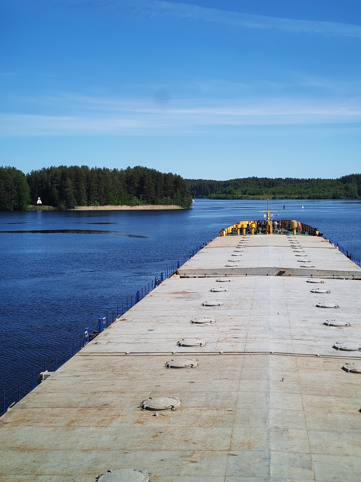 The image presents an expansive view from the deck of a large, flat-topped vessel, possibly a barge or cargo ship, as it navigates a wide, deep blue waterway. The perspective looks aft (towards the stern), where a yellow-painted superstructure is visible, with a small group of people and a flag discernible on it. The vessel's deck, which occupies the foreground and midground, is a weathered gray with numerous evenly spaced circular metal hatches, and is bordered by blue railings.
The waterway is flanked on both sides by dense green forests, primarily coniferous trees. On the left bank, a distinctive white conical structure, likely a navigational marker or small lighthouse, stands out against the dark tree line. Further along the waterway, a white buoy is visible on the water's surface.
The setting suggests a sunny and clear day, likely morning or early afternoon, indicated by the bright blue sky with only a few wispy clouds. The water is calm with gentle ripples, and the overall scene conveys a sense of peaceful transit through a natural landscape. There is no visible text in the image.