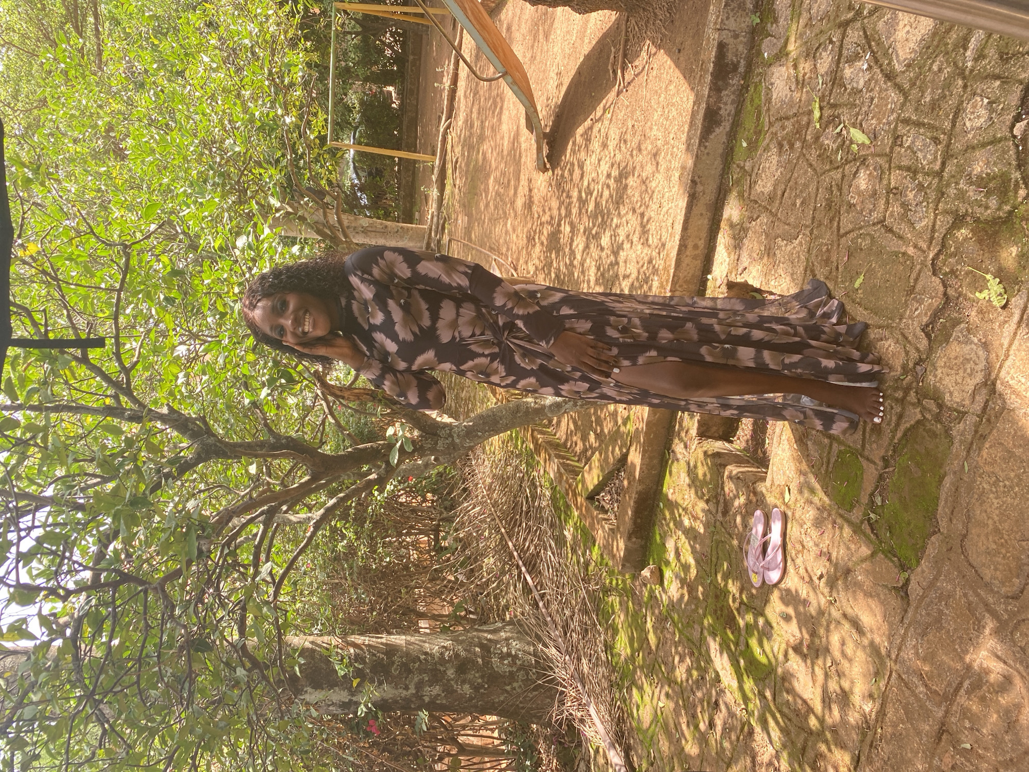 The image depicts a woman standing outdoors on a stone-paved surface, possibly a pathway or patio. She is wearing a long, patterned dress and smiling at the camera. Behind her are trees and greenery, suggesting a garden or park-like setting. A pair of sandals are on the ground near her feet. Based on the information, the location is likely Bukuru, Nigeria.