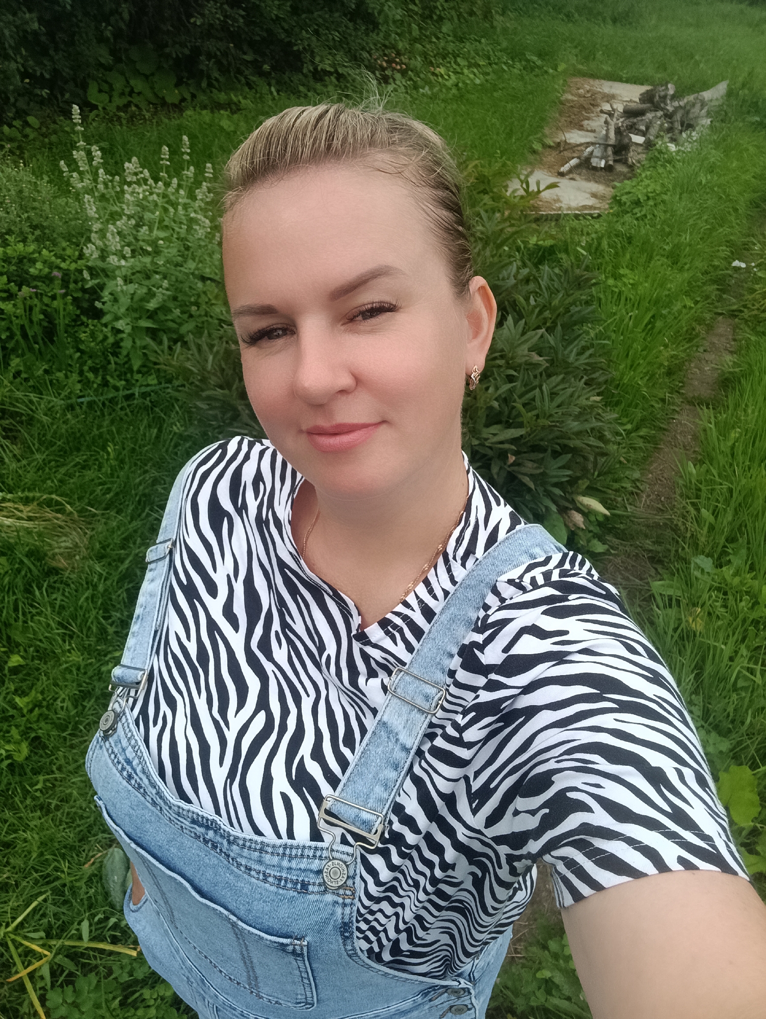 A woman with blonde hair pulled back poses for a selfie outdoors in a grassy area with some green foliage and a dirt path visible. She is wearing a black and white zebra-print shirt and denim overalls. The background shows a patch of light-colored wildflowers and a small area with debris, possibly from construction or landscaping, further back in the image. The lighting suggests it is daytime, with no direct sunlight evident. The location is identified as Chaikovsky, Russia.