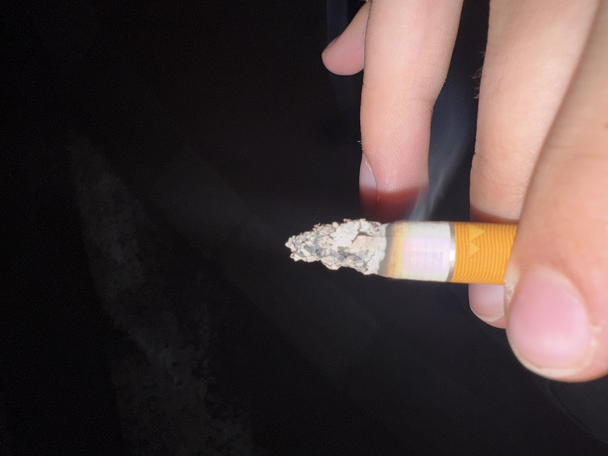 This is a close-up, macro photograph of a lit cigarette being held by a person's hands in an outdoor, dark environment. The cigarette is positioned vertically, with the burning end at the top, showing ash and embers. The filter end of the cigarette is visible towards the bottom, wrapped in orange and white paper. The person's fingers, with visible skin texture and fingernails, are partially obscuring the lower part of the cigarette and the background. The background is predominantly black, with some faint, blurry light or smoke patterns visible, suggesting a night-time or low-light setting. The image composition focuses on the act of holding and potentially smoking the cigarette. There is no visible information in the image that would suggest a specific location within Trenčín, Slovakia.