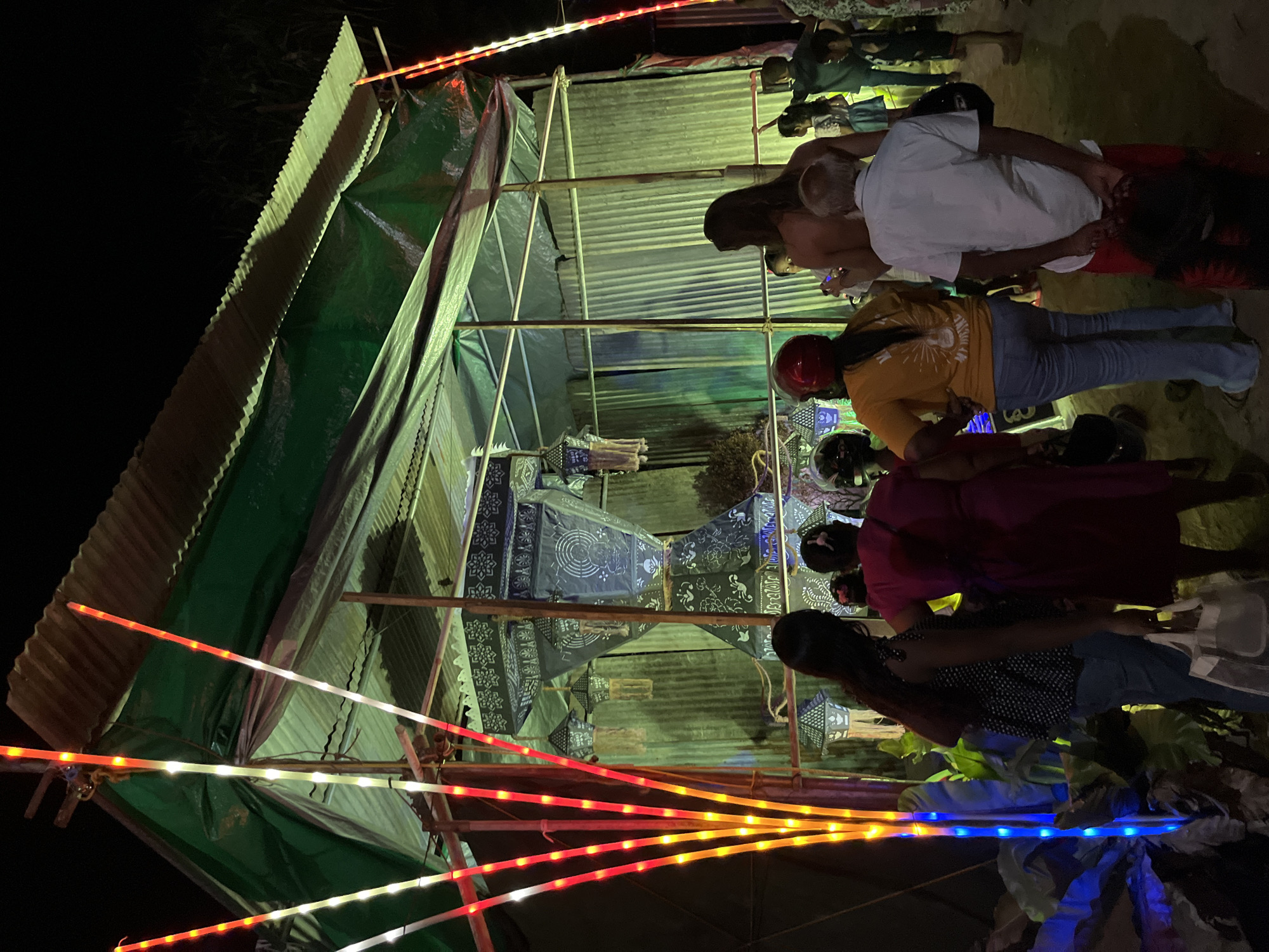 The image depicts an outdoor scene at night, likely a market or a gathering, in Habaraduwa, Sri Lanka. Several people are visible, some standing and others seated or leaning. They are gathered around a temporary structure covered with green tarp and corrugated metal sheeting, which appears to be a stall or a covered area.
The scene is illuminated by strings of colorful LED lights, predominantly red, yellow, and white, which are draped around the structure and extending outwards. These lights create a festive atmosphere. Several lanterns, decorated with intricate patterns in dark blue and white, are hanging within the structure, suggesting a possible cultural event or decoration. Some of the people are wearing casual clothing, and one person is wearing a red helmet.
The ground appears to be unpaved, consistent with an outdoor setting in a tropical location. The overall impression is of a lively nighttime event, possibly a festival or a local market, characterized by the vibrant lighting and the presence of decorative lanterns. The time of day is clearly night, indicated by the darkness surrounding the illuminated area. There is no visible text in the image.