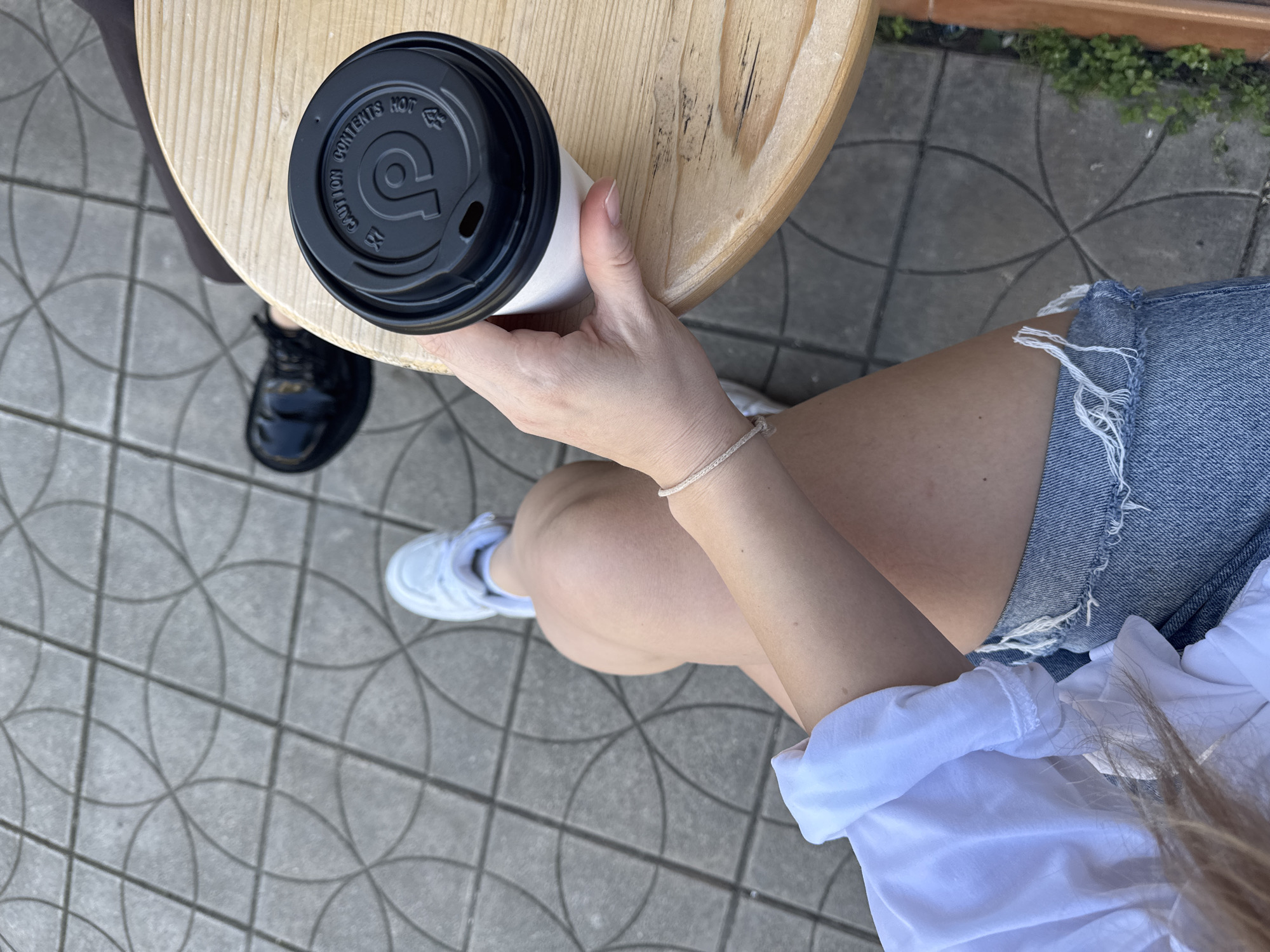 An overhead shot shows a woman holding a white disposable coffee cup with a black lid while sitting at a light-colored wooden table. The table sits on a patterned gray tile surface. The woman wears a white short-sleeved shirt and denim shorts with frayed edges. On her left leg is a delicate bracelet. One white sneaker and one black shoe are visible near the table legs. The scene implies a coffee break, possibly at an outdoor cafe in Bar, Montenegro, indicated by the type of tile and outdoor setting. A bit of greenery appears in the background.