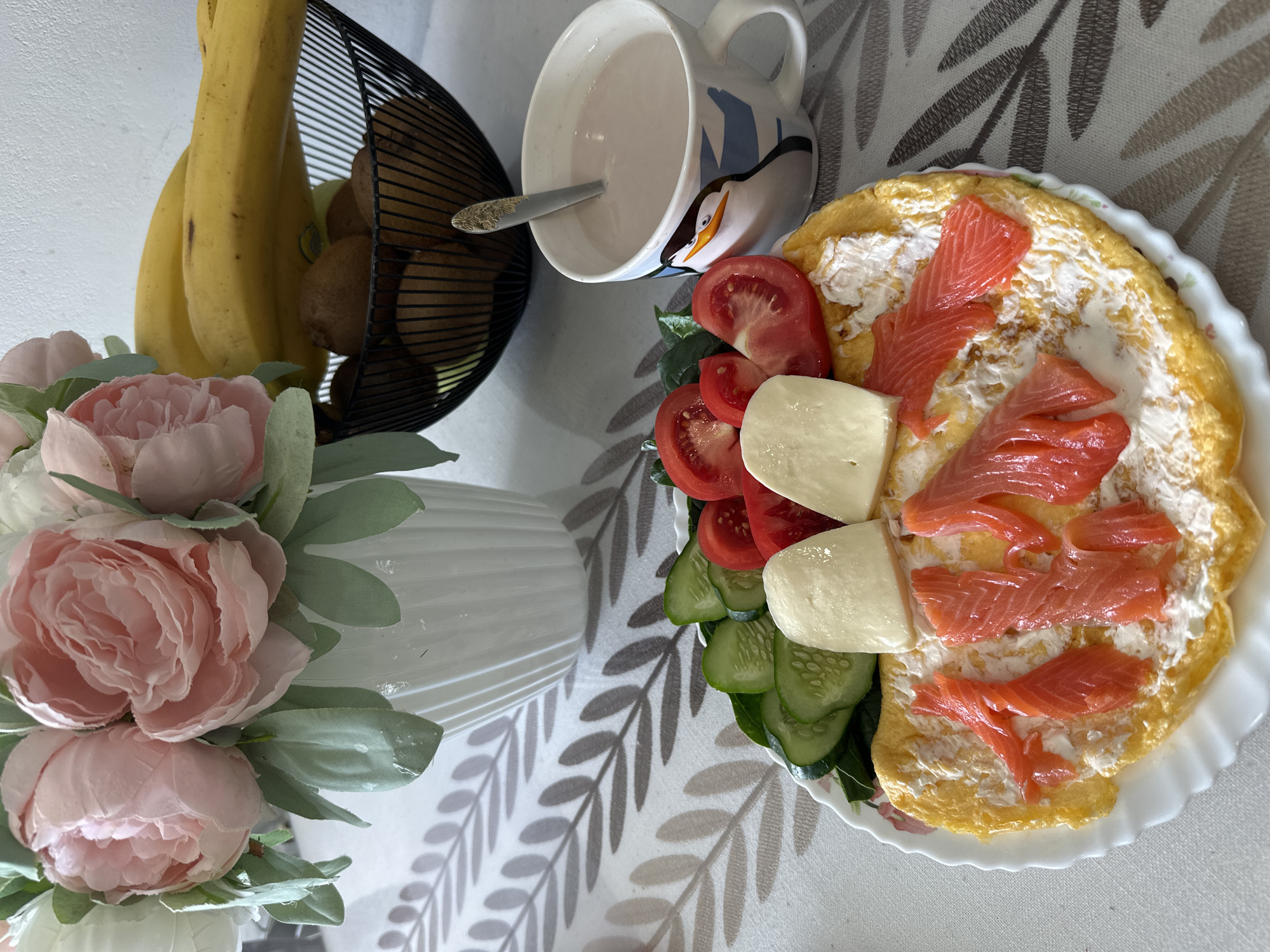 A table is set with a breakfast or brunch arrangement, featuring a plate of omelet topped with cream cheese and salmon, alongside tomato slices, mozzarella cheese, and cucumber slices. A mug containing a liquid and a spoon sits adjacent to the plate. In the background, a wire fruit basket holds bananas and kiwis, while a vase containing pink flowers is also present. The table is covered with a tablecloth patterned with leaf designs. The image does not suggest the city of Sochi or any specific location in Russia.