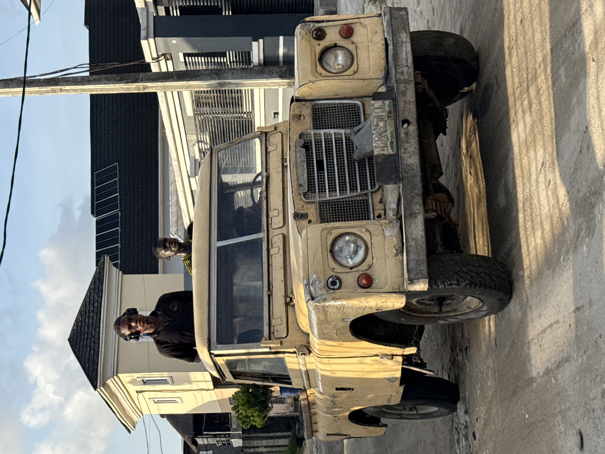 Here's a detailed description of the image:
The image captures a vintage, light-yellow Land Rover vehicle, parked on what appears to be a paved street or road. The vehicle shows signs of age and use. Two men are visible inside the vehicle. One is wearing a black shirt and headphones. The other is wearing a yellow shirt and is positioned behind the first man. The backdrop consists of buildings with modern architectural elements, including what seems to be a rooftop solar panel installation. There is also a utility pole with wires running across the scene. It is most likely located in Ado, Nigeria.