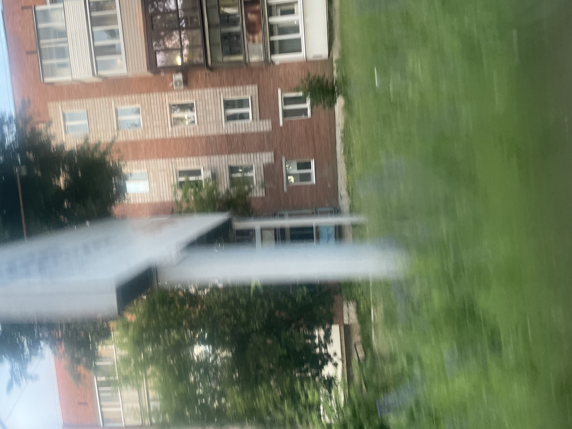 The image shows a blurry, vertically oriented view of an apartment building with a brick facade and several windows. Trees are visible in front of the building, and a grassy area with some white markings is to the right. A light-colored structure, possibly a sign or a roof of a smaller building, is in the foreground. The scene appears to be outdoors, likely in a residential area. The lighting suggests it might be daytime, but the blurriness makes it difficult to determine the exact time or weather conditions. The overall impression is that of a slightly disheveled, everyday urban or suburban environment in Votkinsk, Russia. No people are clearly identifiable.