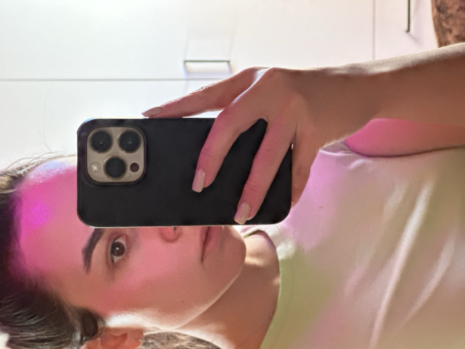 An indoor mirror selfie depicts an adult female subject in the city of 3-Y Uchastok, Russia. The subject's face, partially obscured, is illuminated from the left by a strong pink/magenta light source, casting a noticeable hue across her forehead and the visible right side of her face. Her right eye is open, looking directly towards the mirror. She has dark hair and fair skin. Her right hand, with fingernails painted a light neutral color, holds a dark-colored smartphone, equipped with a triple camera system, positioned centrally in front of her face. She is dressed in a light-colored, likely light green, crew-neck t-shirt. The background is a plain white surface, possibly a wall or wardrobe, with a subtle handle visible on the right side.