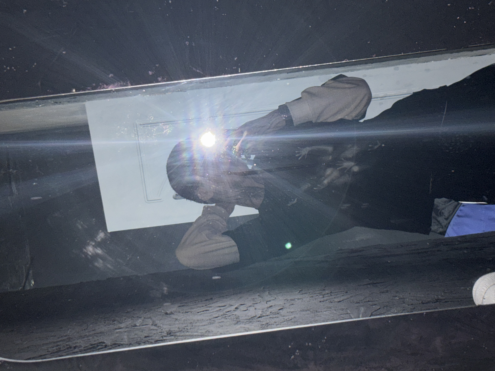 The image shows a person in a dark environment, possibly underground. The individual is wearing light-colored sleeves and holding a bright light source, creating a glare. The reflection suggests the scene is taking place within a structure. The floor appears to be covered in dark, possibly gritty material. A blue object is also visible in the background. The location in the city of Saratov, Russia is unknown.