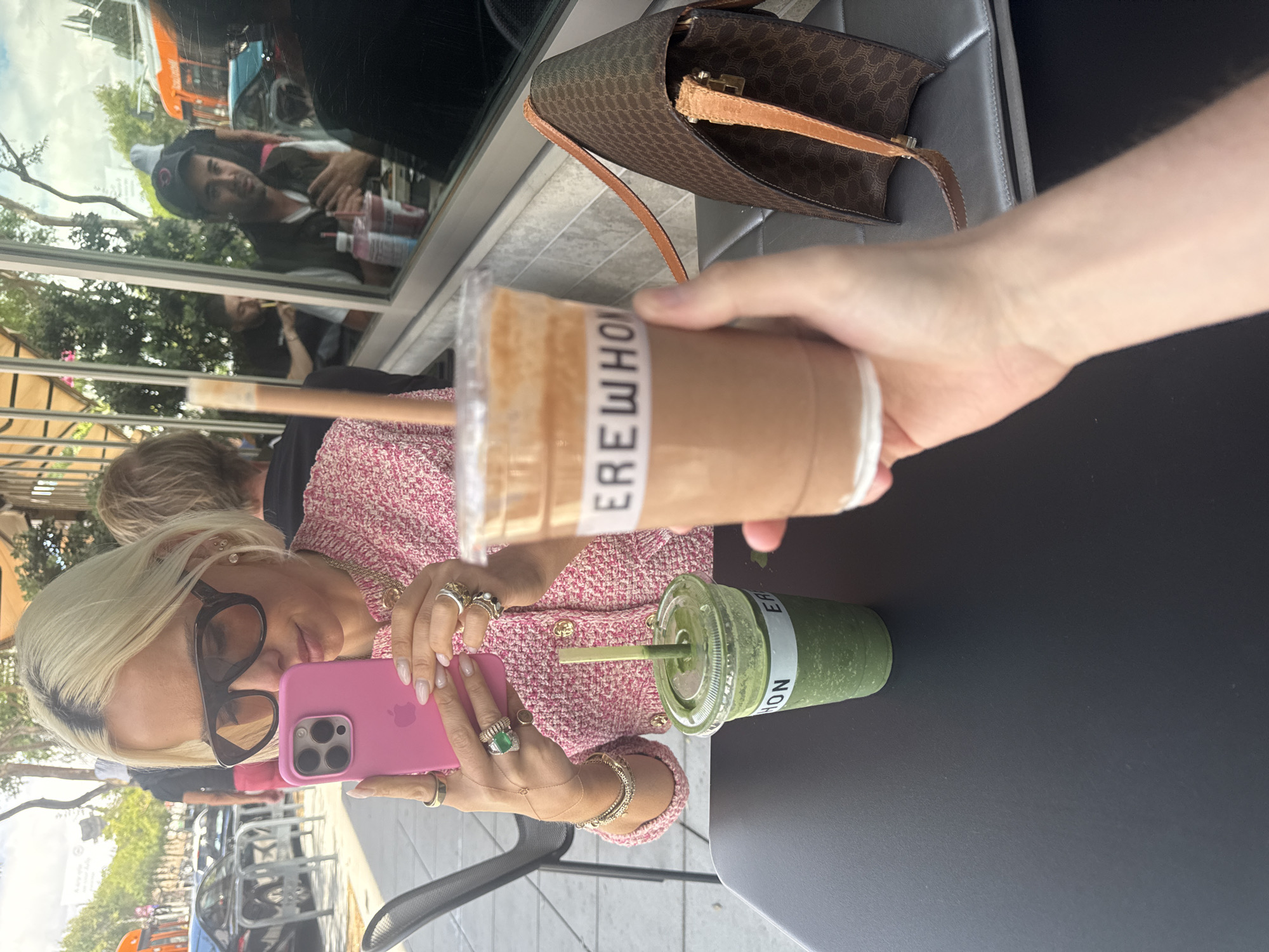 The image captures a scene in West Hollywood, USA, centered around a woman taking a photo with her pink smartphone. She has blonde hair, wears sunglasses, and a pink knit cardigan. She is adorned with multiple rings and bracelets. She holds her phone in her right hand and appears to be taking a picture of a hand holding an Erewhon branded drink above a table with a green drink of the same brand on it. On the table, there is also a brown Celine handbag. In the background, a window reflects passersby and the city environment. The scene suggests an outdoor cafe setting.