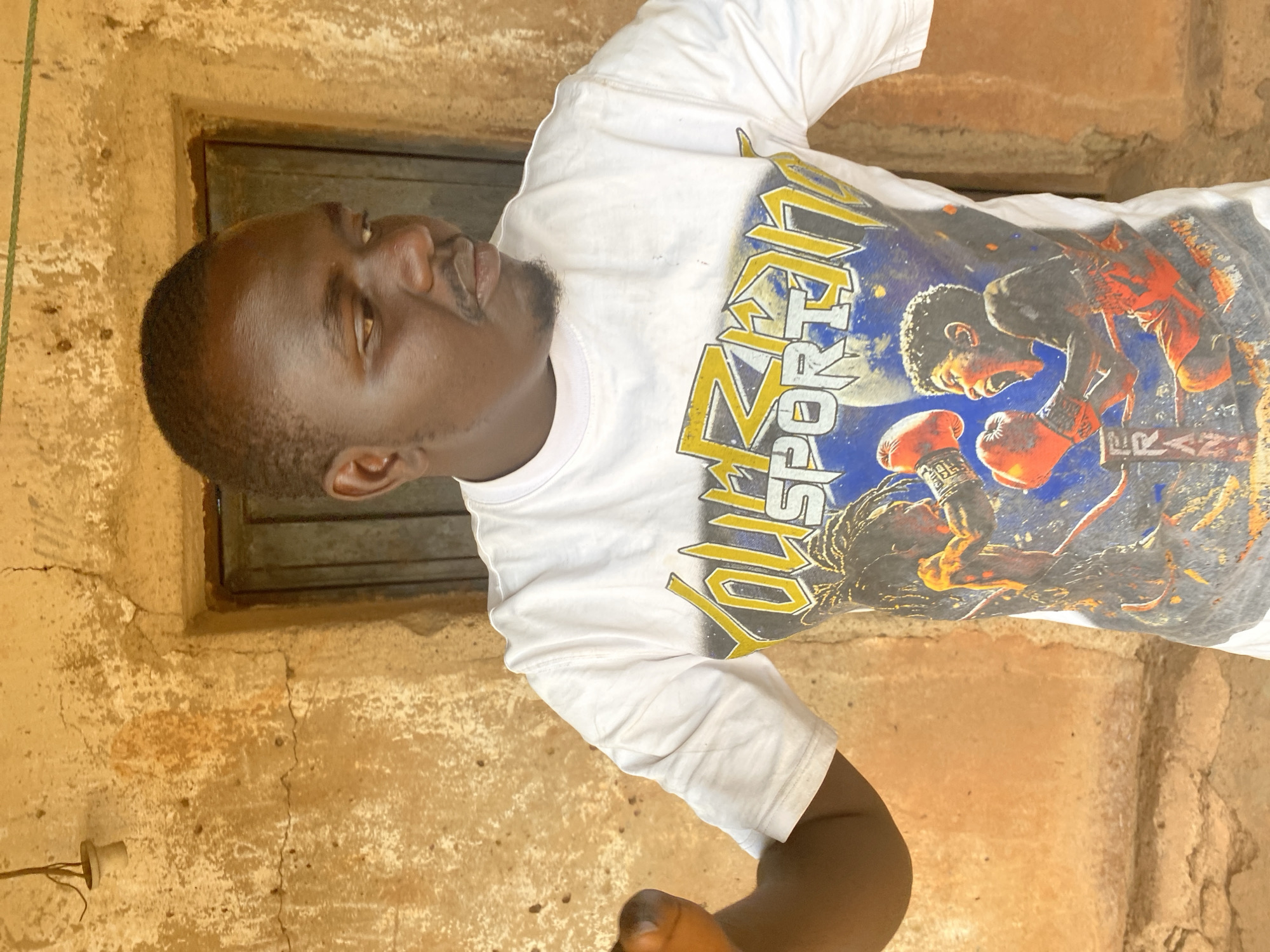 The image, taken in Bukuru, Nigeria, features a young man of dark complexion captured in a relaxed pose outdoors during the daytime. He has short hair and a neatly trimmed beard, looking slightly upwards and to the right with a neutral expression. He is wearing a white short-sleeved T-shirt adorned with a vibrant graphic print. The print prominently displays the words "YOU SPORT" in large yellow letters, alongside an illustration of two boxers in action, one wearing red gloves, and the word "FRAN" visible beneath one of the figures.
The background consists of a textured, earthy-toned wall, likely made of mud or concrete, exhibiting cracks and areas of discoloration, indicative of an older structure. Behind the man's head, there is a dark, rectangular opening, possibly a window or a recessed part of the wall. A small, white electrical fitting, resembling a bare bulb holder with wires, is visible on the lower left portion of the wall. The bright, natural lighting suggests the photo was taken during daylight hours.