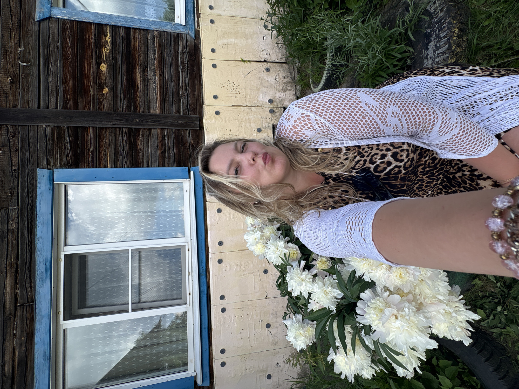 A woman is taking a selfie in front of a wooden house with blue trim around the windows. She has blonde hair and is wearing a leopard print dress and a white crocheted cardigan. In the foreground, there is a large bouquet of white peonies. The house appears to be in a rural or suburban setting, possibly in Chaikovsky, Russia. The wall of the house is covered in light-colored panels with dark circular fastenings. The ground is covered in grass and some vegetation.
