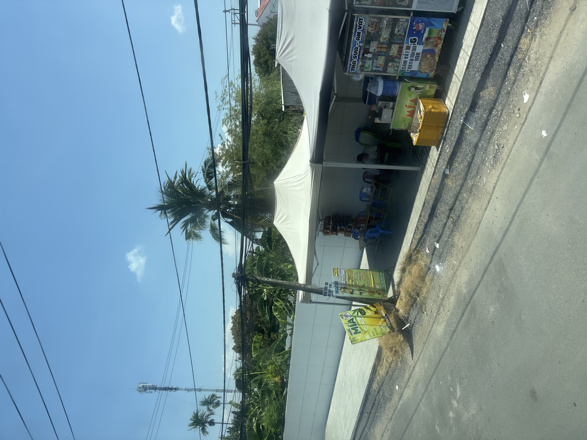 The image captures a street scene under a clear blue sky with some scattered clouds. A prominent cell tower stands tall on the left side of the frame, with several palm trees visible around it. Overhead, a tangle of power lines crisscrosses the sky. In the foreground, a paved road runs parallel to a sidewalk. Along the sidewalk, a small roadside stall operates, covered by white canopies. The stall displays various colorful signs, advertising drinks and food items, with some products visible on shelves. A few plastic chairs are set up near the stall, suggesting a waiting or seating area. Behind the stall, a white wall encloses a lush green area with dense foliage. The overall scene depicts a typical day in a developing urban or semi-urban environment, likely in a tropical region.
