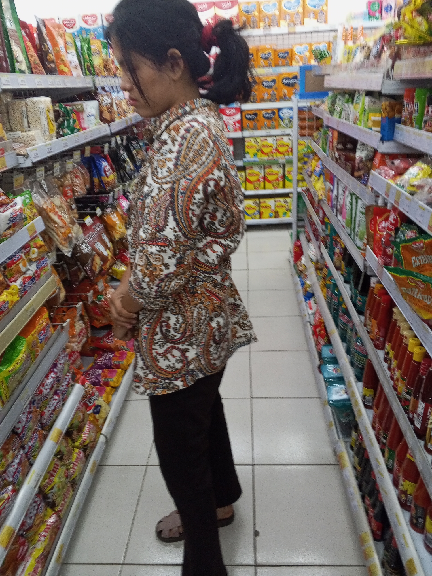 A woman with dark hair pulled back in a ponytail stands in a grocery store aisle, facing away from the camera. She is wearing a patterned shirt with paisley designs in white, brown, and orange, and dark pants. Her hands are clasped in front of her. She is looking towards shelves stocked with various food products. The shelves on the left are filled with bagged snacks and other packaged goods, while the shelves on the right are lined with bottles, possibly condiments or sauces. The store appears to be well-lit with fluorescent lighting. The floor is tiled in a light color. The scene is consistent with a grocery store environment in Ciruas, Indonesia.