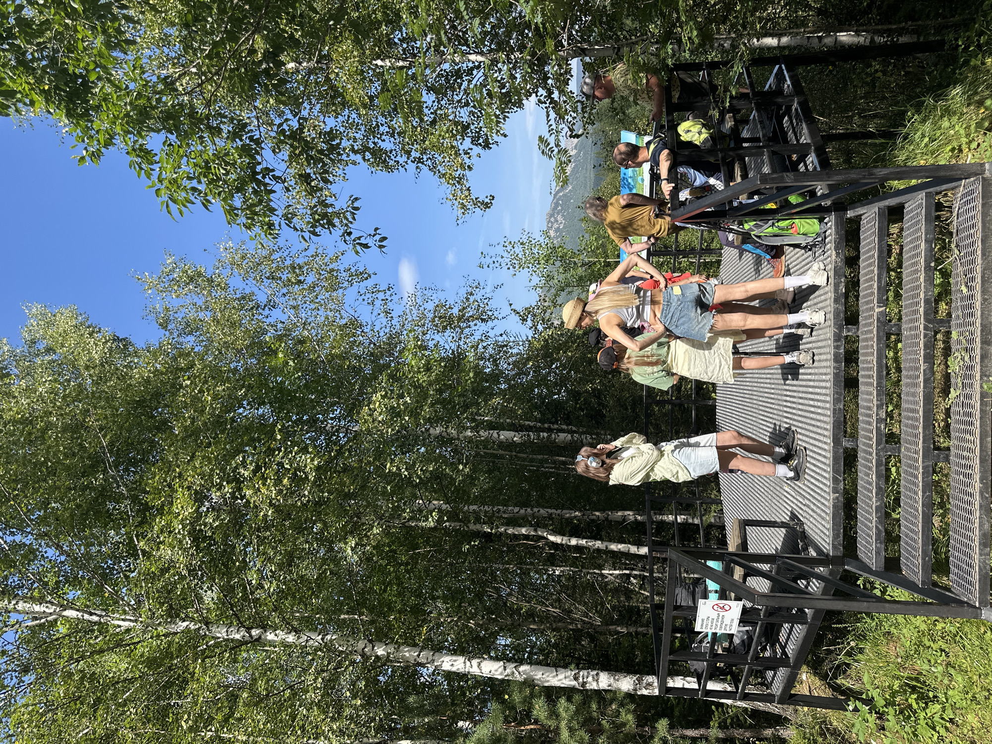 A group of people stand on a metal observation deck in a forest, looking out at a scenic mountain view. The deck is elevated and accessed by stairs. Several people are visible, including a woman in a straw hat and denim shorts, a woman in a light green shirt and shorts, and a man in a military-style shirt. A sign with Cyrillic text and a prohibition symbol is attached to the railing. The surrounding environment is a dense forest with birch trees and undergrowth. The sky is clear and blue with a few scattered clouds. The overall scene suggests an outdoor recreational area or a tourist viewpoint.