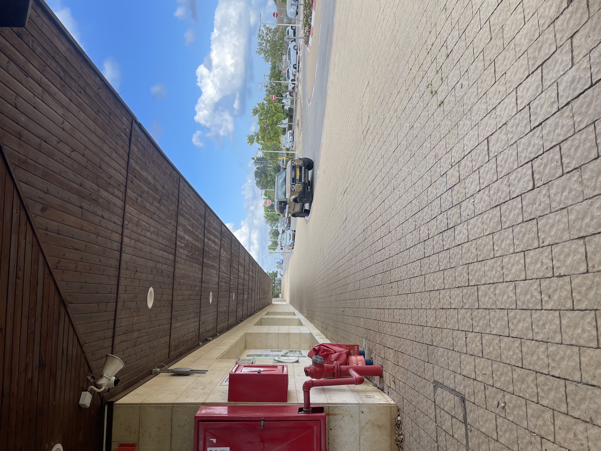 The image depicts a street scene in Even Yehuda, Israel, characterized by a covered walkway with a wooden ceiling and recessed lighting, adjacent to a paved sidewalk. On the left side of the walkway, a red fire extinguisher cabinet is mounted on a stone-colored building, with a red fire hydrant and its associated piping in front of it. The sidewalk is made of light-colored rectangular pavers. Beyond the sidewalk, a road is visible with several cars, including a light brown SUV in the foreground and other vehicles further down the road. The sky is blue with scattered white clouds. The scene appears to be a public area, possibly in front of a commercial building or a community center.