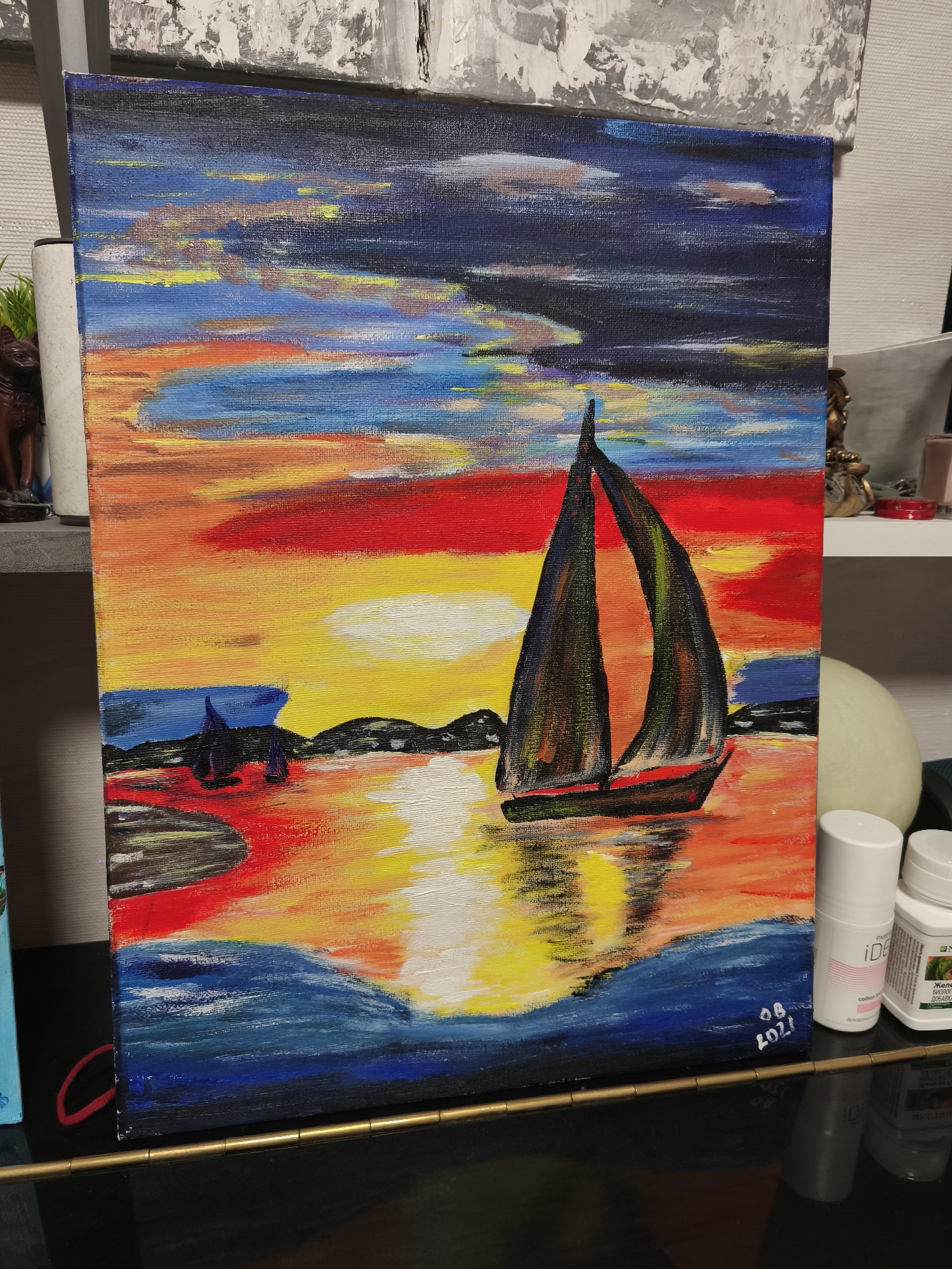 An acrylic painting, depicting a sailboat on water during a vibrant sunset or sunrise, is prominently displayed. The sky is a dramatic blend of deep blues, reds, oranges, and yellows, with some clouds illuminated by the setting/rising sun. The central sun casts a bright, reflective path of white and yellow light across the water. The main sailboat is a dark silhouette with two sails, positioned on the right side of the painting, its reflection visible in the water. To the left, smaller, darker boats are visible near a distant, silhouetted landmass. The water transitions from fiery reds and oranges near the center to deep blues in the foreground.
The painting features visible brushstrokes, giving it a textured appearance. In the bottom right corner, a signature "08 2021" is inscribed, indicating the date.
The painting is positioned upright, leaning against a surface, with various objects visible around it. To the left, a dark, carved figure and a white cylindrical object are partially seen. Above and behind the painting, another abstract artwork with grey and white textures is visible. To the right, on a shelf, are two white bottles: one is a deodorant labeled "iDE..." and "comfort," and the other is a supplement bottle with Cyrillic text, including "БИОЛОГИЧЕСКИ АКТИВНАЯ ДОБАВКА" (Biologically Active Supplement) and "ЖЕЛ...". A spherical object and a metallic decorative item are also present. A metallic, segmented rod runs horizontally across the bottom of the frame.
