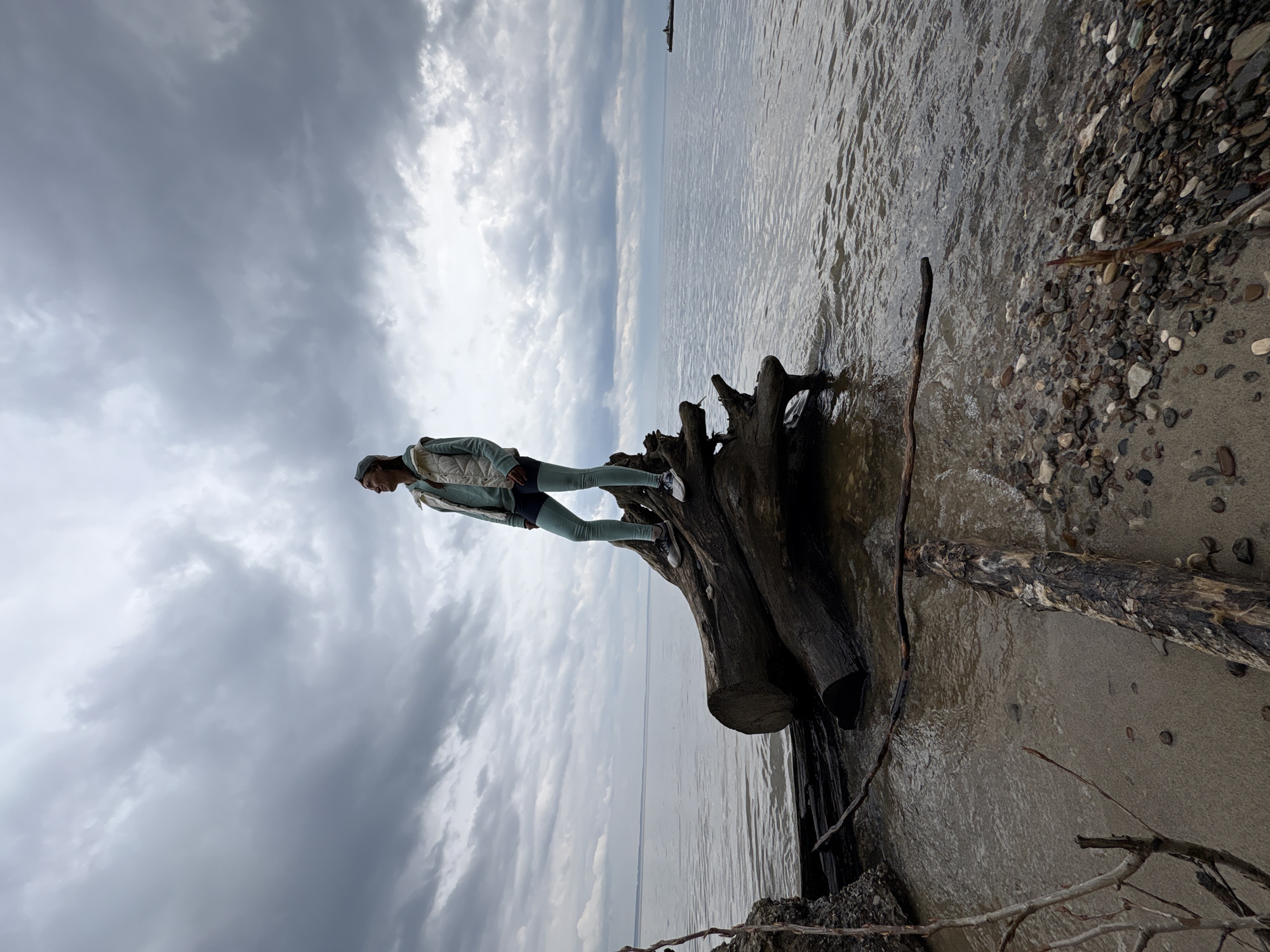 A person stands on a large, waterlogged tree trunk on a beach. The water meets a grey, cloudy sky at the horizon. To the right of the tree trunk is a beach of sand and rocks. The person is wearing a light jacket, dark pants, and light-colored shoes. The image appears to be rotated, possibly to create an effect of disorientation or imbalance. The location is a beach in Ulyanovsk, Russia.