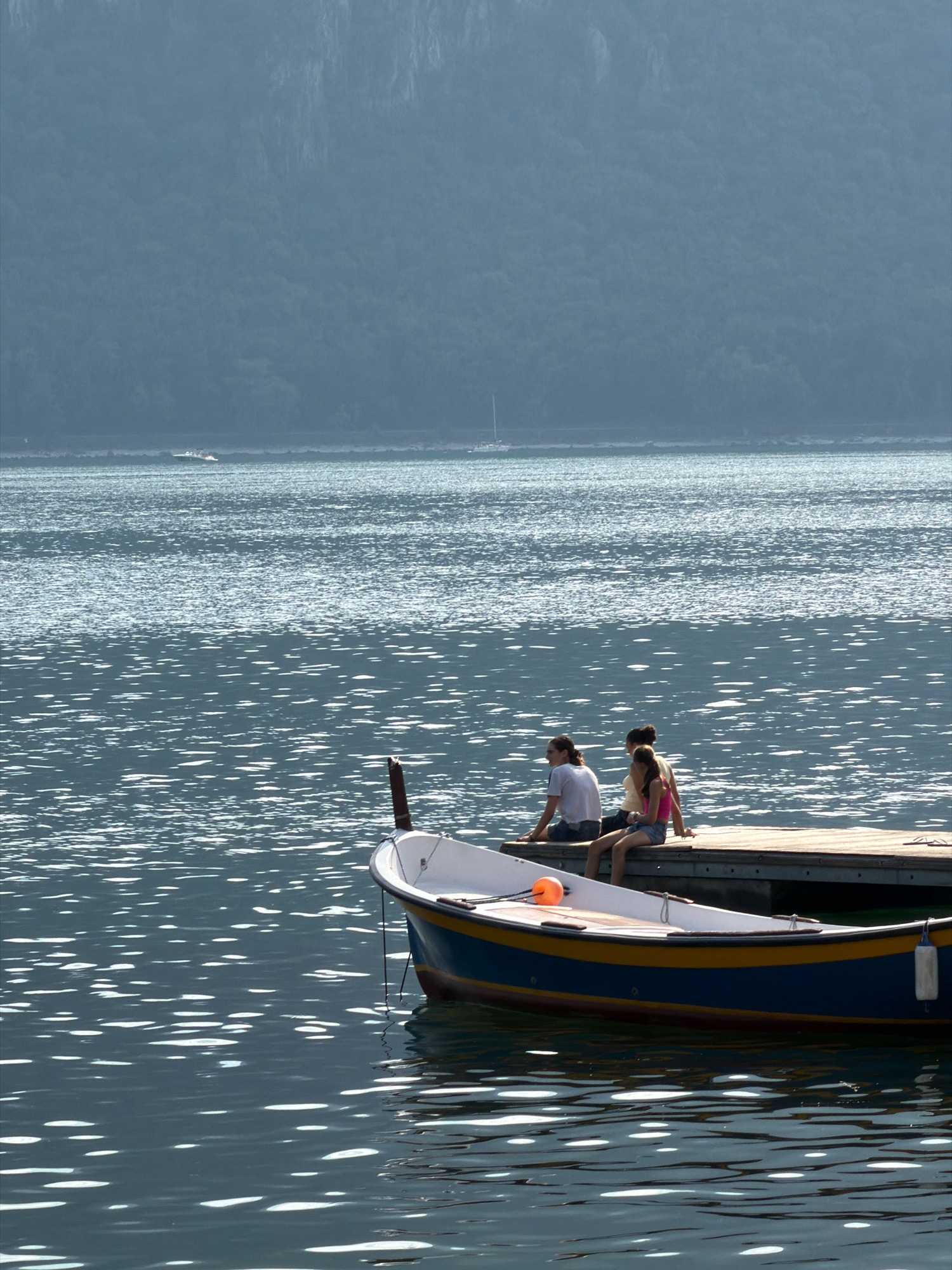 This is a vertical, eye-level shot of a serene lake, likely Lake Como, with a misty mountain range in the background. In the foreground, a small, wooden rowboat with a blue and yellow hull is docked. Three individuals, two women and a person whose gender is not discernible, are seated on the edge of a wooden pier adjacent to the boat. The person on the far left is wearing a white t-shirt and dark shorts, while the person in the middle wears a yellow top and shorts. The person on the far right is wearing a pink tank top and denim shorts. The water's surface is dappled with sunlight, creating a shimmering effect. The overall atmosphere is peaceful and idyllic, suggesting a relaxed summer day in a location like Mandello del Lario, Italy, known for its picturesque lake and mountain scenery.