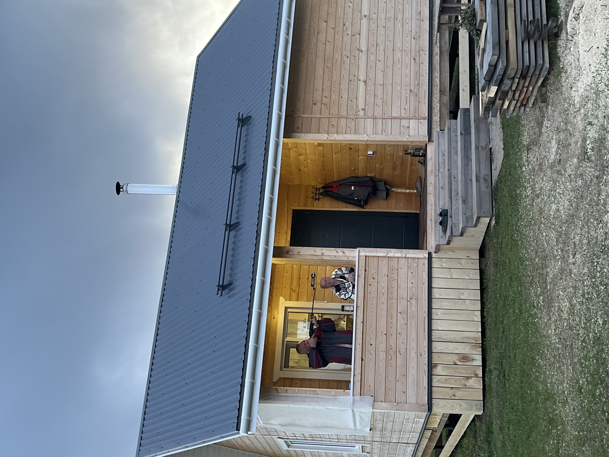 A daytime shot depicts a wooden house with a gray metal roof, a chimney, and a porch where two men are standing. The house appears to be located in a rural or suburban setting, with grass visible on the right. One man on the porch is wearing a dark jacket and holding what appears to be a selfie stick. The other man is wearing a patterned sweater. A black coat is hanging on the wall near the doorway. The location is presumed to be Гаревая, Russia.