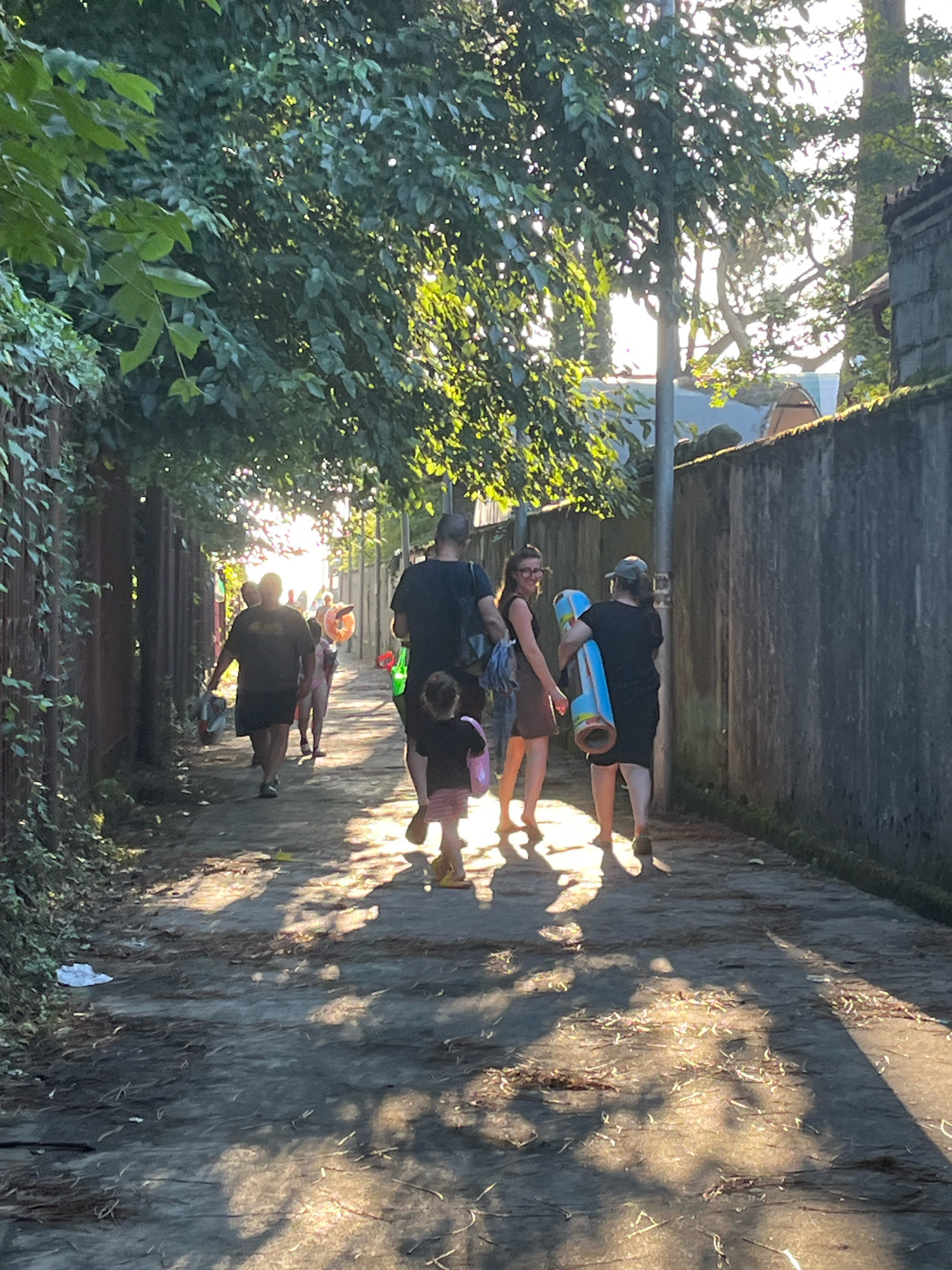 The image depicts a pathway in Ureki, Georgia, characterized by dappled sunlight filtering through trees lining the left side. A grey concrete wall runs along the right side of the path. Several individuals are walking away from the viewer down the path. In the foreground, a young child in a pink skirt and dark top walks ahead, holding a pink bag. Behind the child, a group of adults follows, some carrying inflatable items and bags. One adult on the right carries a large blue inflatable tube. The path itself is paved with a mixture of dirt and gravel. The overall atmosphere suggests a casual outing, possibly to the beach, given the presence of beach-related items.