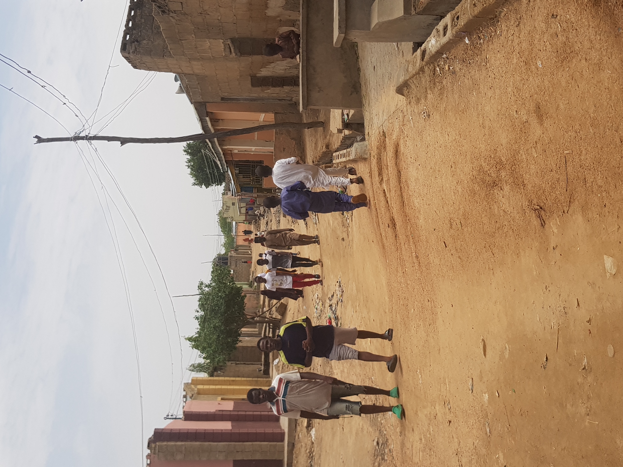 A street in Dutsinma B, Nigeria, shows a group of people walking along a dirt road flanked by buildings. The building on the left is a pinkish hue, and another building is on the right. Overhead, an unadorned utility pole with wires. Two men stand in the foreground. The first one is wearing a white shirt with a blue stripe and gray shorts. The second one is wearing a black shirt with yellow stripes and gray shorts.