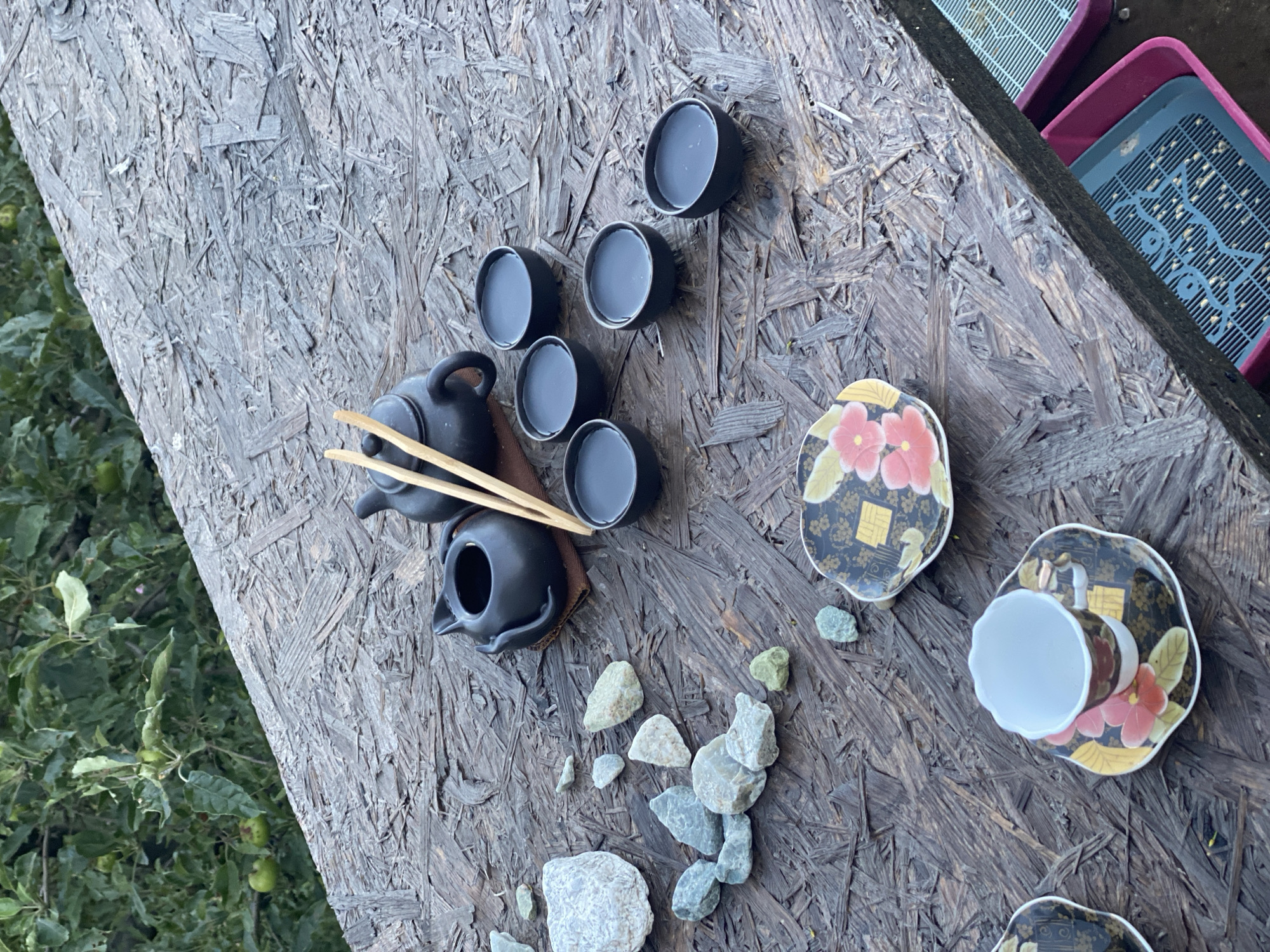 A still life image features a tea set on a rustic wooden surface. The tea set consists of a small black teapot with a lid, a smaller black pot, and several small black cups filled with a dark liquid. Two wooden chopsticks rest on the teapot. Scattered around the tea set are various stones of different sizes and colors. In the foreground, two teacups with saucers adorned with floral patterns are visible. The background shows green foliage, suggesting an outdoor setting. The surface appears to be made of particleboard. The image is taken from a slightly elevated angle.