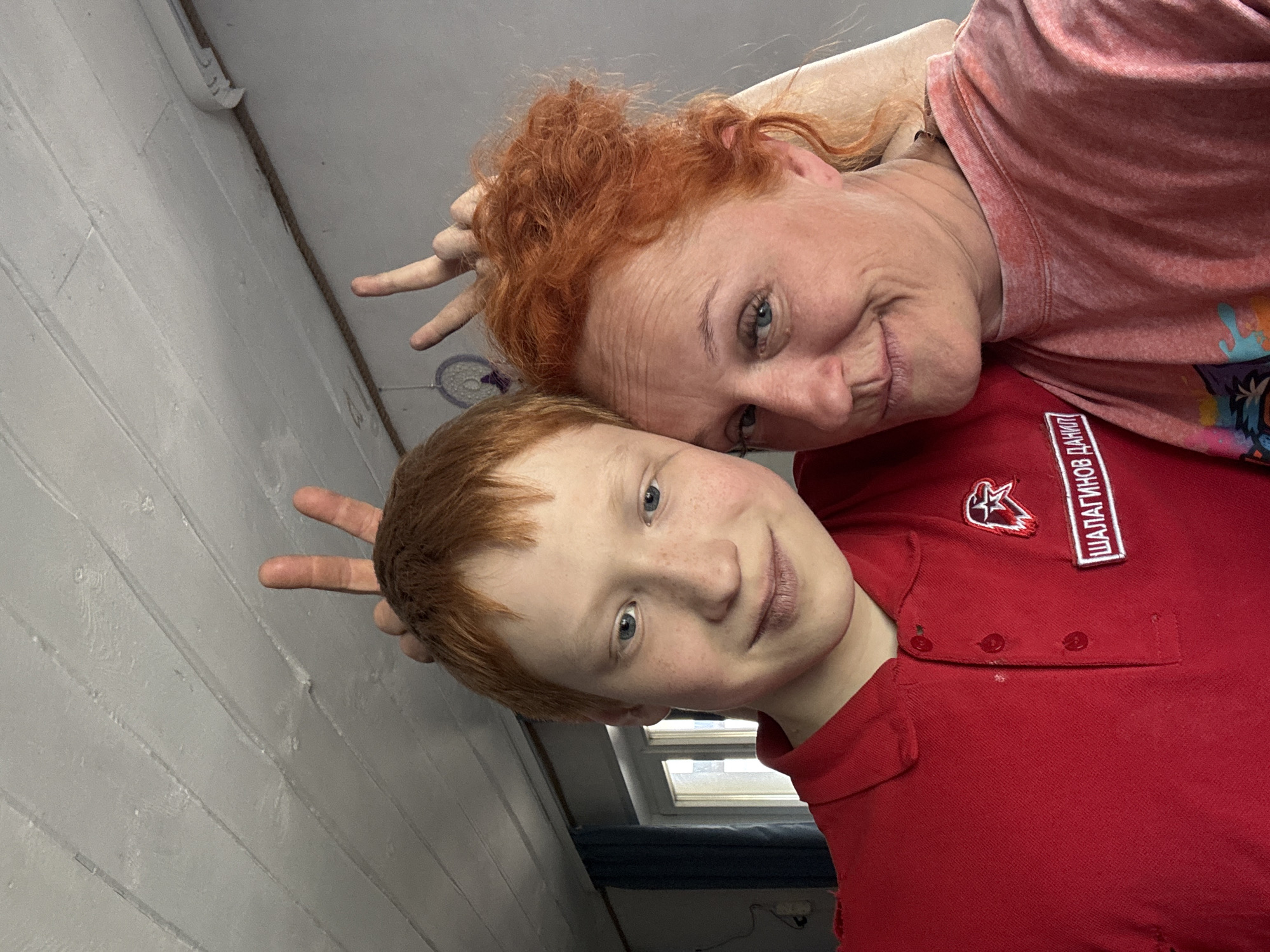 A selfie-style photograph shows a woman and a boy with red hair posing playfully indoors. The woman, with her head resting on the boy's, makes bunny ears with her fingers behind his head. The boy similarly poses with his fingers behind the woman's head. The woman is wearing a light red shirt and the boy is wearing a red polo shirt with a patch on the left side. The text on the patch is in Cyrillic: "ШАЛАГИНОВ ДАНИЛ". The location is likely Chaikovsky, Russia, based on the text in the patch. The background shows a painted, paneled wall, and a window can be seen partially behind the boy.
