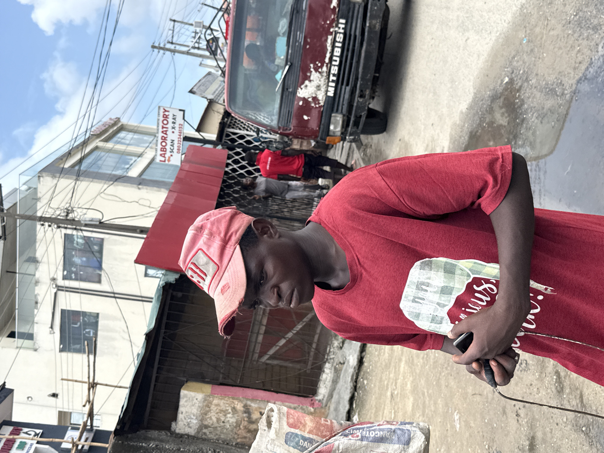 A medium outdoor shot depicts a young black man in Ado, Nigeria, standing on a street. He is wearing a red t-shirt and a red baseball cap, and he is looking down at a smartphone he holds in his hands. An electrical cord is attached to the phone. Behind him is a red Mitsubishi truck. To the left, a building has a sign reading "LABORATORY * SCAN * X-RAY 08032346152." Other people are visible in the background near the building. Bags of cement and debris litter the foreground. Overhead are power lines. The weather appears overcast.