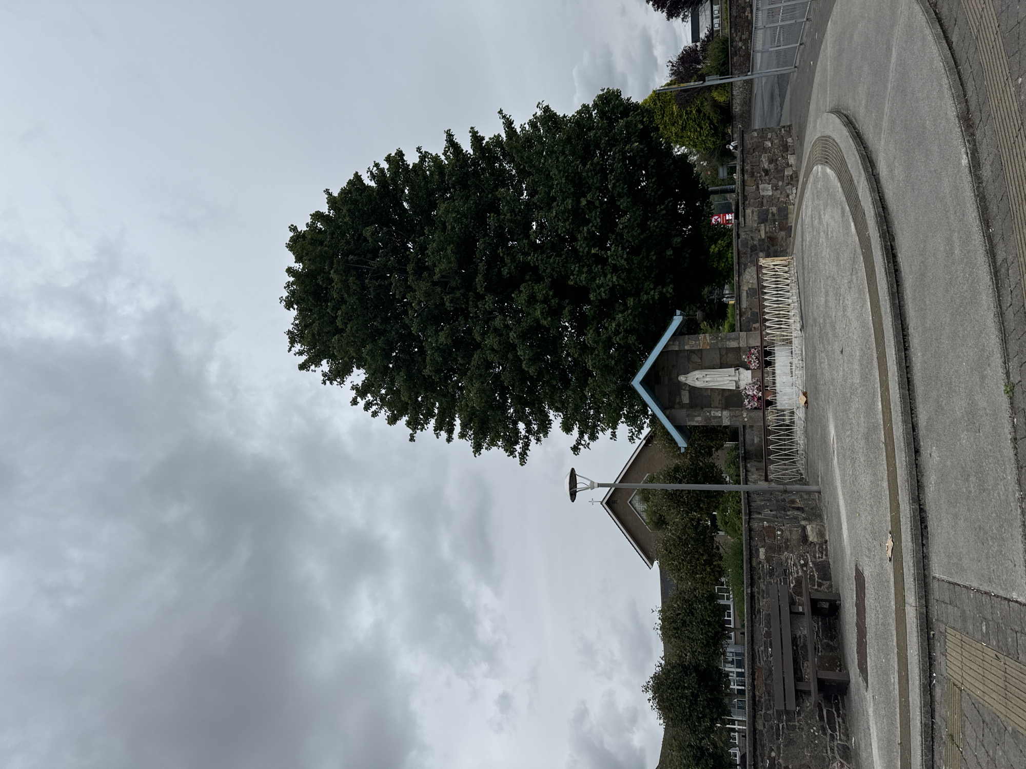 The image depicts an outdoor scene under an overcast sky, likely in Waterford, Ireland. In the foreground, a curved concrete pathway with tactile paving leads to a small shrine. The shrine features a statue of the Virgin Mary under a blue-roofed structure with stone walls. A white picket fence surrounds the immediate area of the shrine. To the left of the shrine, a wooden bench sits on a stone wall. Behind the wall, a large, mature tree dominates the mid-ground. Further back, the roof and upper portion of a building with a cross on its gable are visible, suggesting a religious institution like a church or school. The background includes more trees and a street with a faint glimpse of a building. The scene is quiet and appears to be devoid of any immediate human activity.