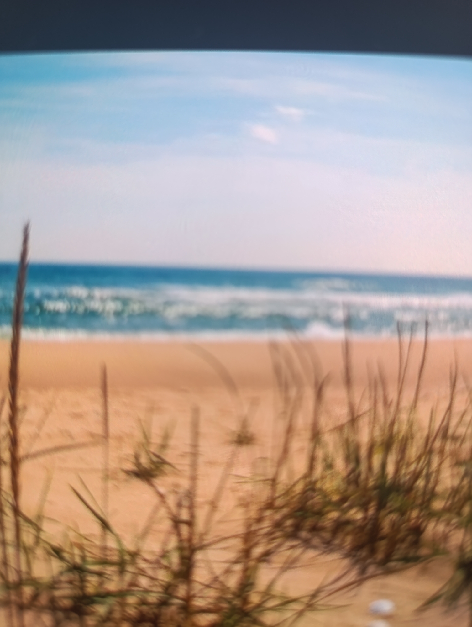 The image depicts a serene beach scene. In the foreground, tall blades of grass are blurred and out of focus, suggesting they are close to the viewer. Behind the grass, a sandy beach stretches towards the ocean. The ocean is a vibrant blue with white waves crashing gently on the shore. The sky above is a soft blue with hints of white clouds. The overall impression is one of natural beauty and tranquility. The location is Unknown, Unknown.