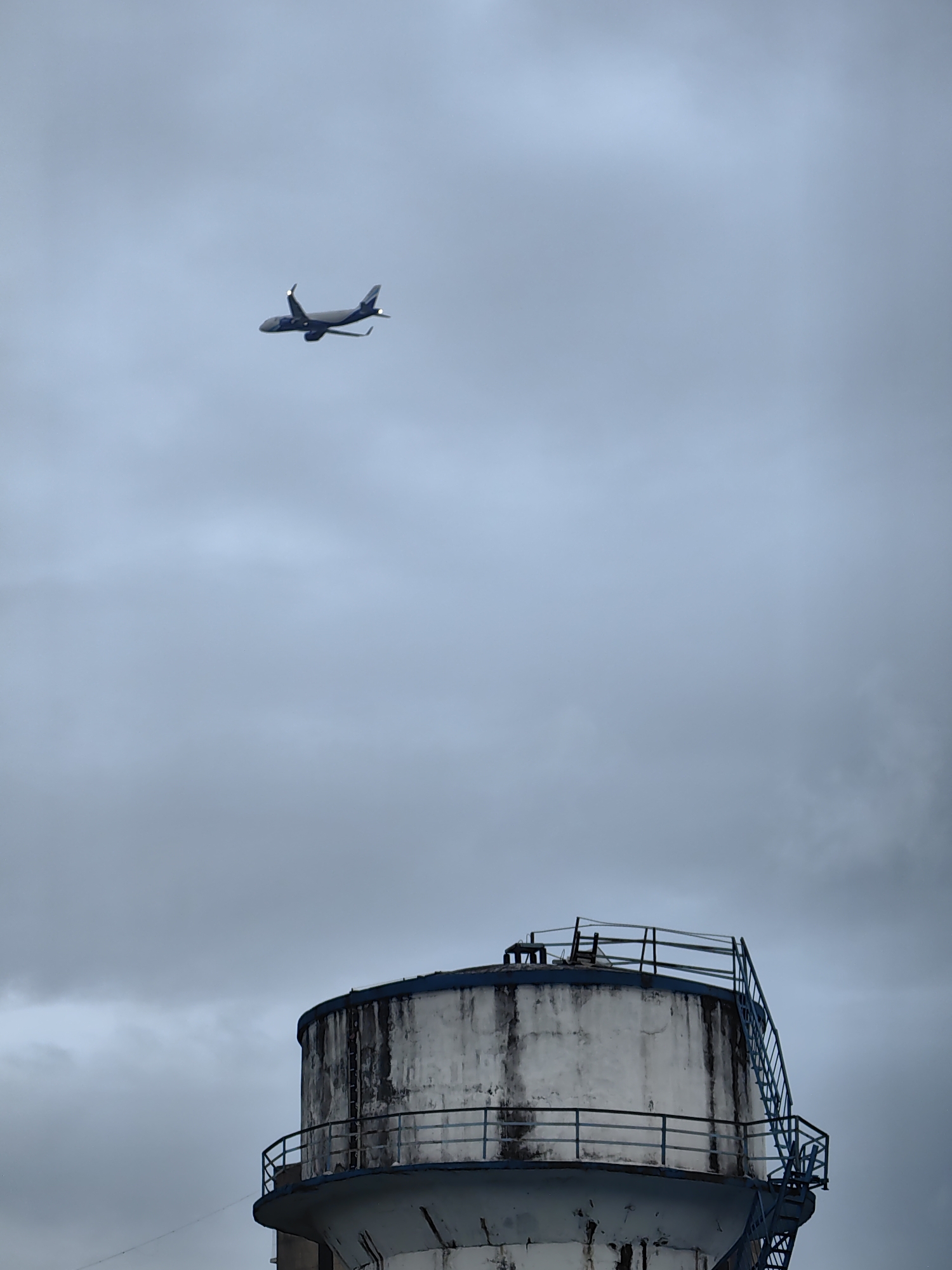 An airplane flies in a cloudy sky above a water tower in Mumbai, India. The airplane is a commercial jet, painted dark blue and white, with its landing gear down. It is flying from left to right across the frame. The water tower is a large, cylindrical structure with a weathered white exterior and a blue railing around its top. A metal staircase with blue railings ascends the side of the tower. The sky is overcast and a dull gray, with patches of lighter clouds.