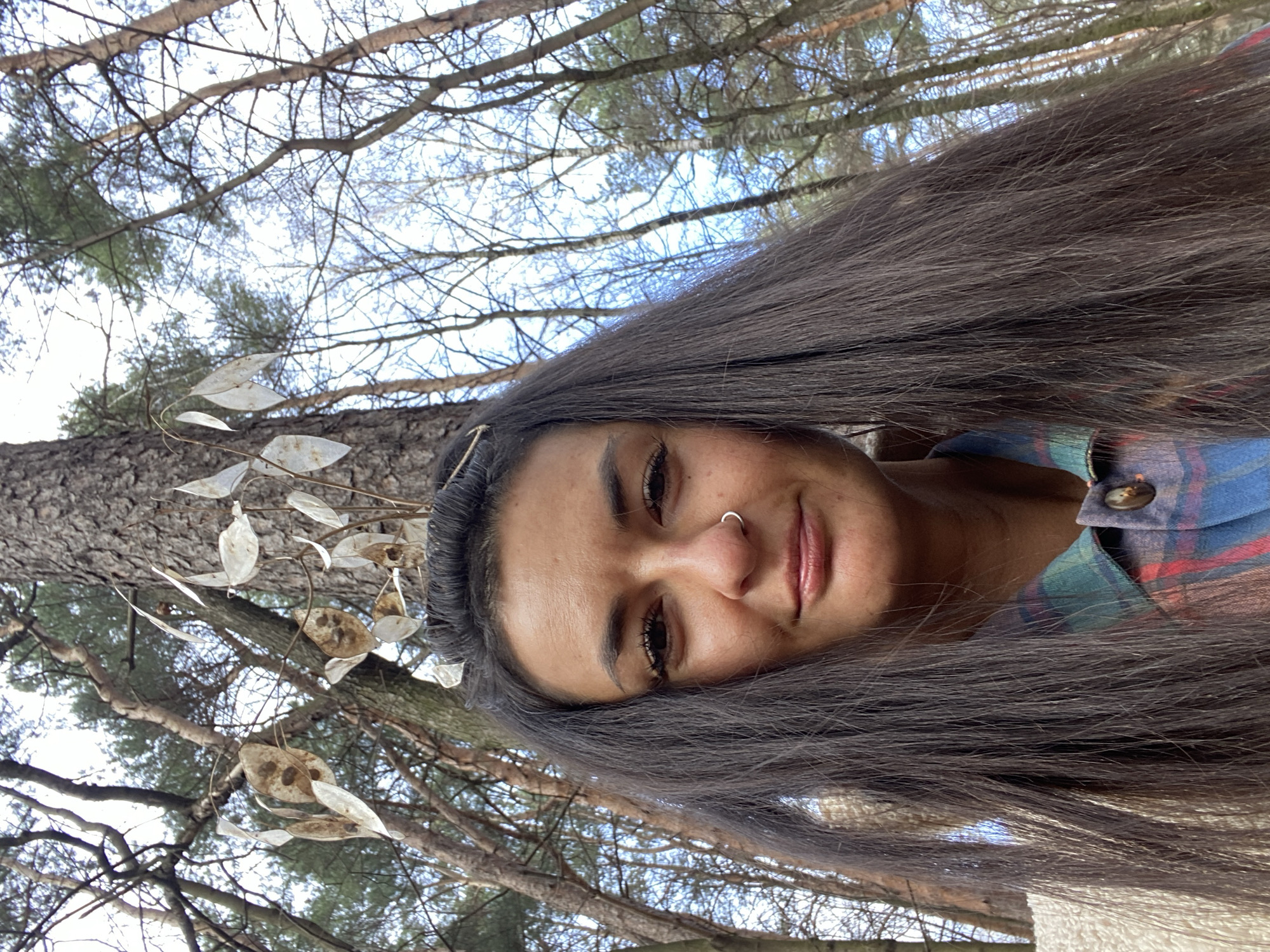 A portrait-style image features a woman captured from the chest up, centered against a natural backdrop. She possesses long, dark hair, a faint smile, and a silver hoop nose ring on her left nostril. She is wearing a blue, red, and green plaid shirt, with a cream-colored, textured outer garment visible on her left shoulder. Several dried, translucent plant seed pods are positioned decoratively within her hair above her forehead. The background consists of a dense forest environment, prominently featuring the rough bark of a large pine tree directly behind her head. Numerous bare deciduous trees and other conifers rise towards an overcast or brightly illuminated sky. This wooded setting is located within a forest park in Moscow, Russia.