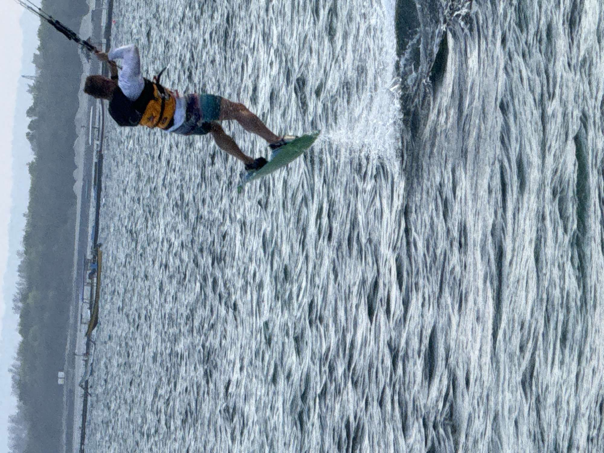 A male kitesurfer is captured mid-air over choppy, dark blue water. He is wearing a white long-sleeved shirt, an orange life vest, and patterned shorts. He holds the kite lines above his head, controlling a light blue kite. His green kiteboard is angled beneath him, with spray erupting from the water's surface where he has just launched. In the background, a treeline borders the horizon, with faint structures suggesting a coastline. The water displays numerous small waves and ripples, indicating movement and activity in the sea. This activity is likely occurring off the coast of Denpasar, Indonesia, a region known for its watersports.